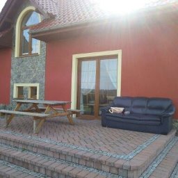 Firma Budowlana Kos-Bud Krzysztof Kosowski - Czerwony dom jednorodzinny z kamienną elewacją, drewnianym stołem piknikowym i czarną skórzaną sofą na brukowanym tarasie.