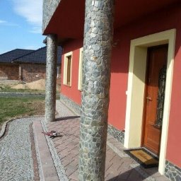 Firma Budowlana Kos-Bud Krzysztof Kosowski - Wykończenie elewacji domu z użyciem kamienia naturalnego na kolumnach i cokole, widoczna również kostka brukowa i narzędzie szlifierskie.