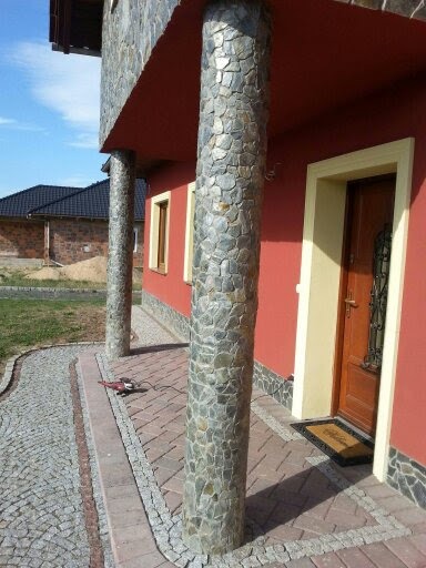 Wykończenie elewacji domu z użyciem kamienia naturalnego na kolumnach i cokole, widoczna również kostka brukowa i narzędzie szlifierskie.