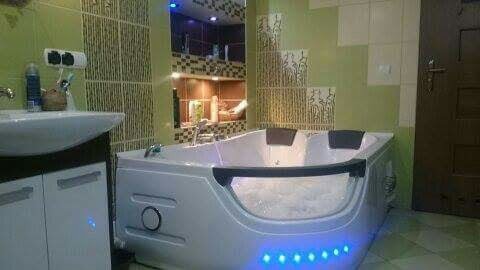 Nowoczesna wanna z hydromasażem i niebieskim podświetleniem LED w łazience z zielonymi ścianami i mozaikową zabudową wnęki.