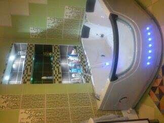 Nowoczesna wanna narożna z hydromasażem i oświetleniem LED w łazience z zielono-beżowymi płytkami i wnęką z półkami na kosmetyki.