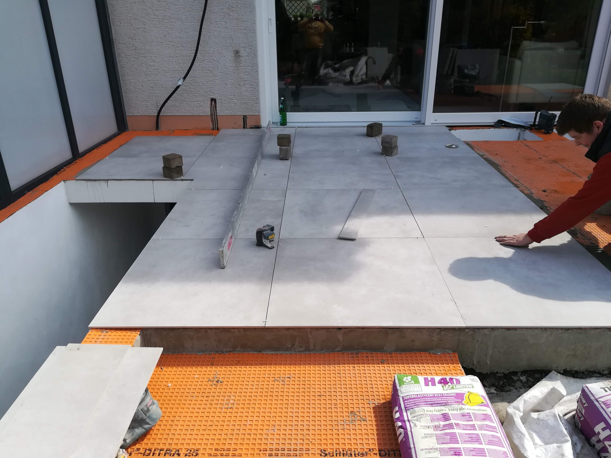 Układanie jasnoszarych płytek na pomarańczowej membranie izolacyjnej na tarasie, widoczne narzędzia, poziomica, miarka, worki z klejem i pracownik dociskający płytkę.