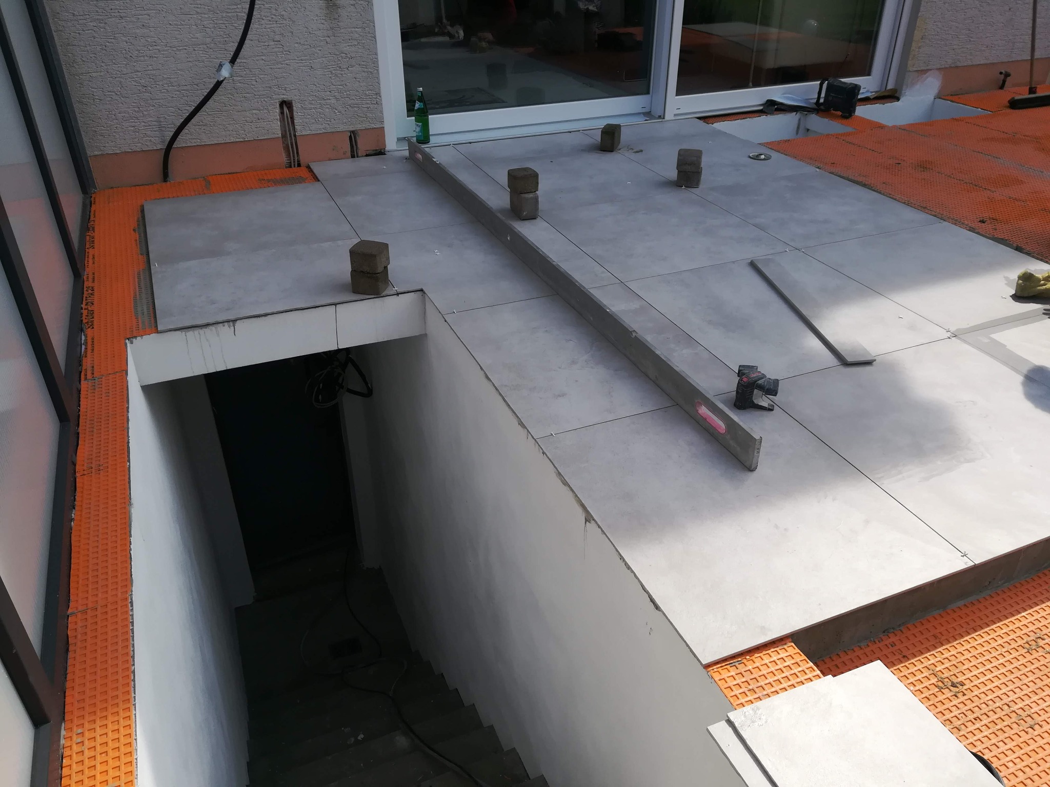 Układanie szarych płytek na tarasie przy wejściu do piwnicy, widoczna poziomica i kostki brukowe jako podpory, pomarańczowa mata drenażowa pod płytkami.