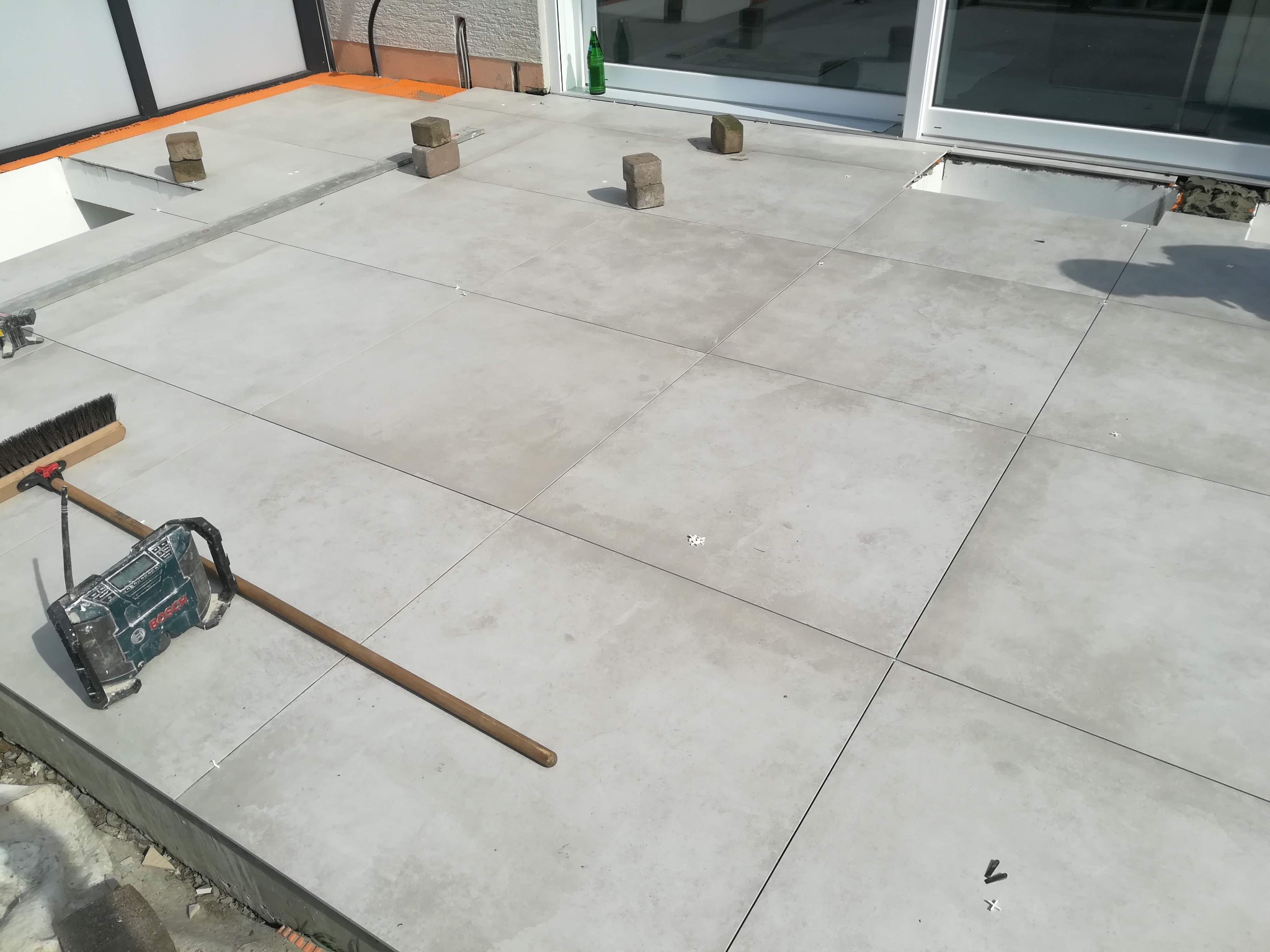Montaż szarych płytek na tarasie zewnętrznym, widoczny laser krzyżowy Bosch, drewniane klocki dystansowe oraz szczotka do zamiatania, częściowo ukończona powierzchnia.