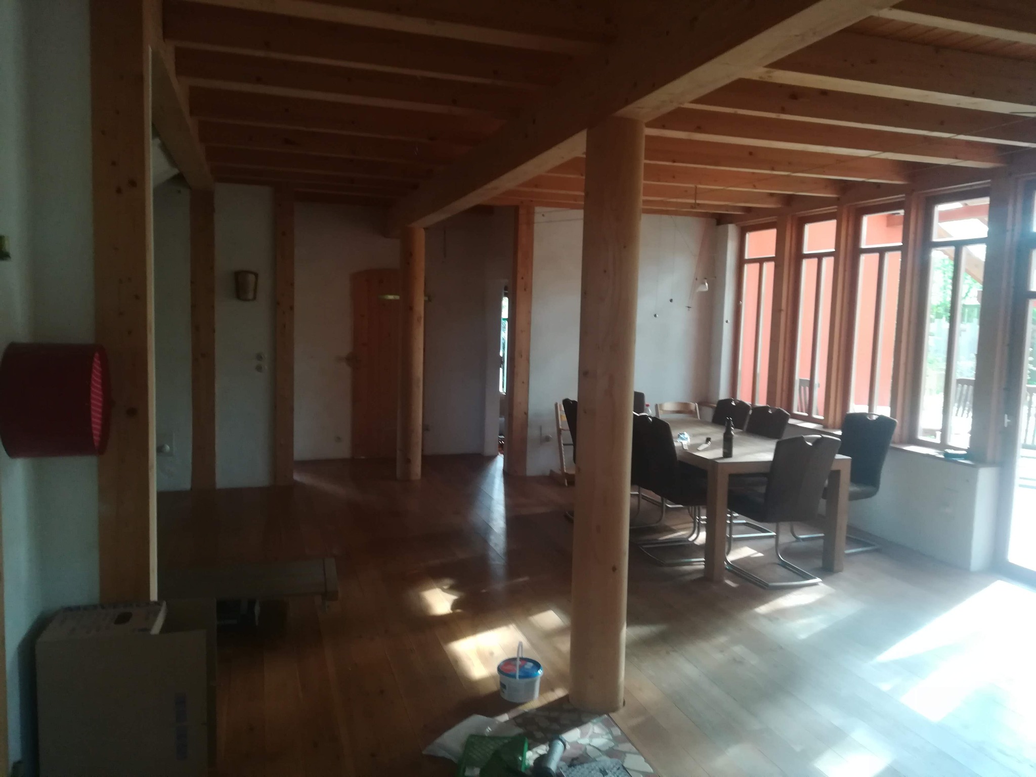 Przestronne wnętrze w trakcie wykończenia, widoczne drewniane elementy konstrukcyjne, podłoga z desek, stół z krzesłami przy oknie, kartony i wiadro z farbą w rogu pomieszczenia.