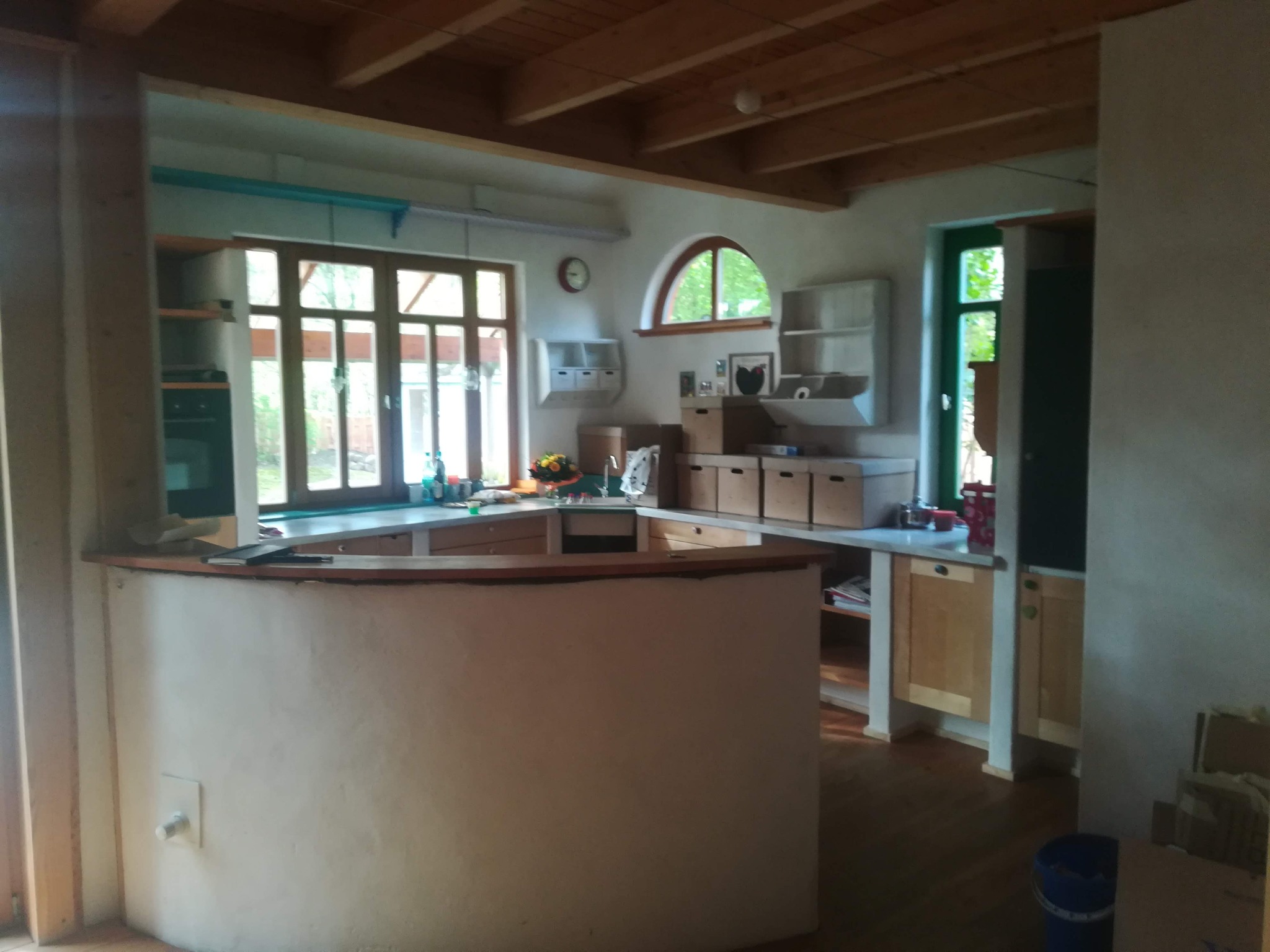 Jasna kuchnia z drewnianym sufitem i oknami, widoczny półwysep kuchenny z blatem, skrzynki kartonowe na blacie, wnętrze w trakcie wykończenia.