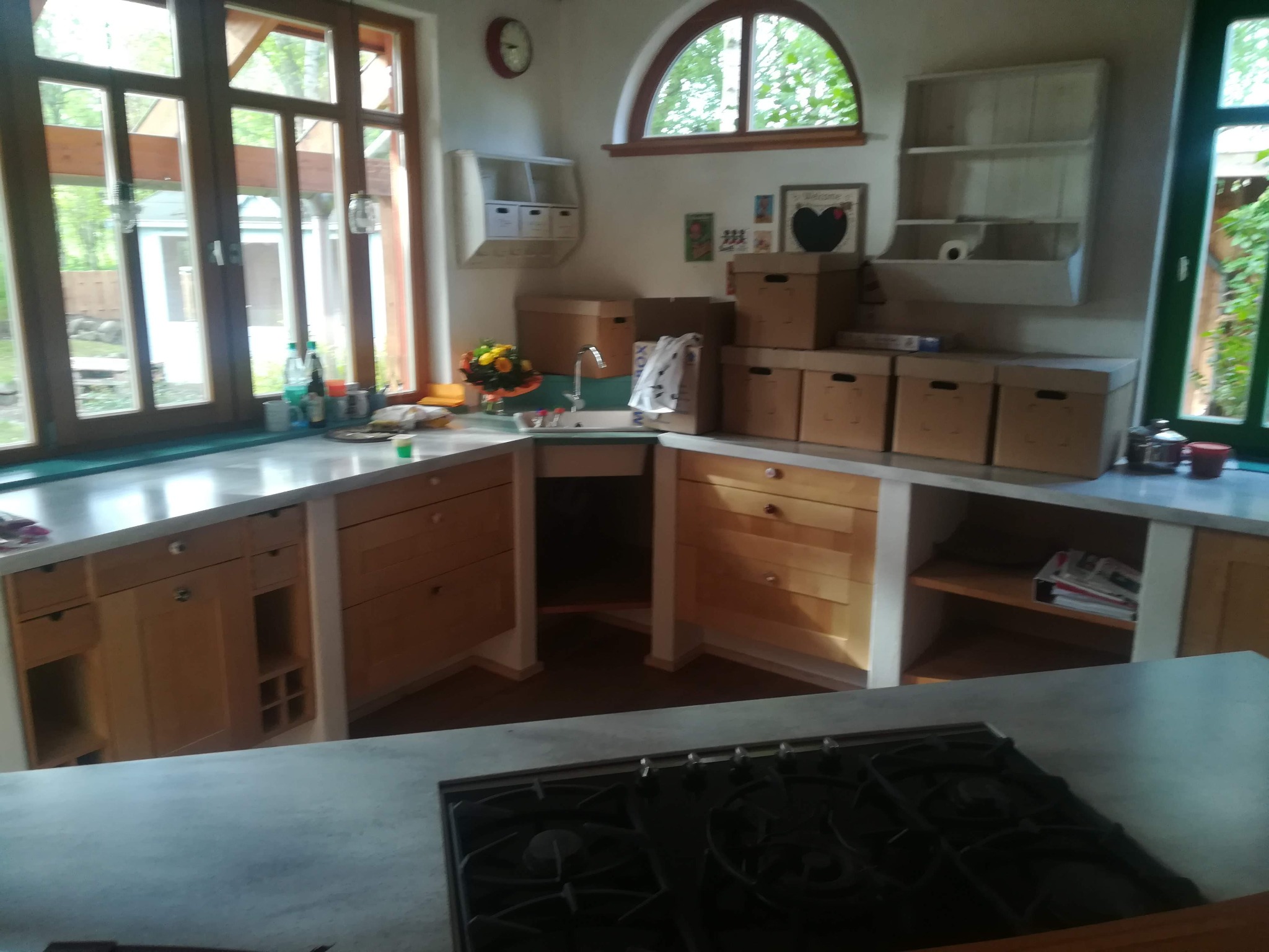 Jasna kuchnia z drewnianymi szafkami i szarym blatem roboczym, widoczne kartonowe pudła na blacie, okno z widokiem na zieleń, płyta gazowa na pierwszym planie.