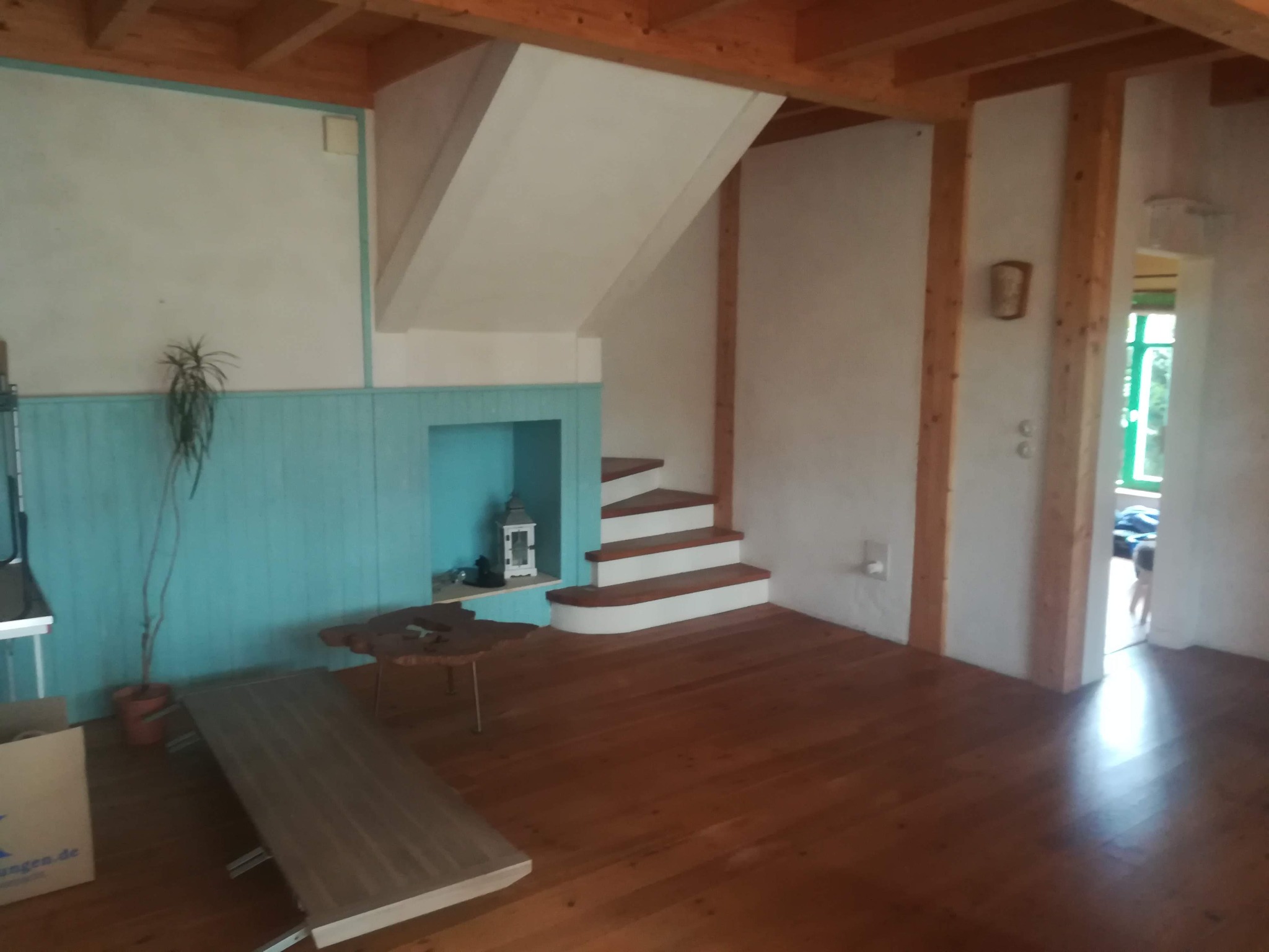 Surowe drewno na podłodze i belkach stropowych w pomieszczeniu z niebieską boazerią i białymi ścianami, schody z drewnianymi stopniami i białą obudową, okno z zieloną ramą w oddali.