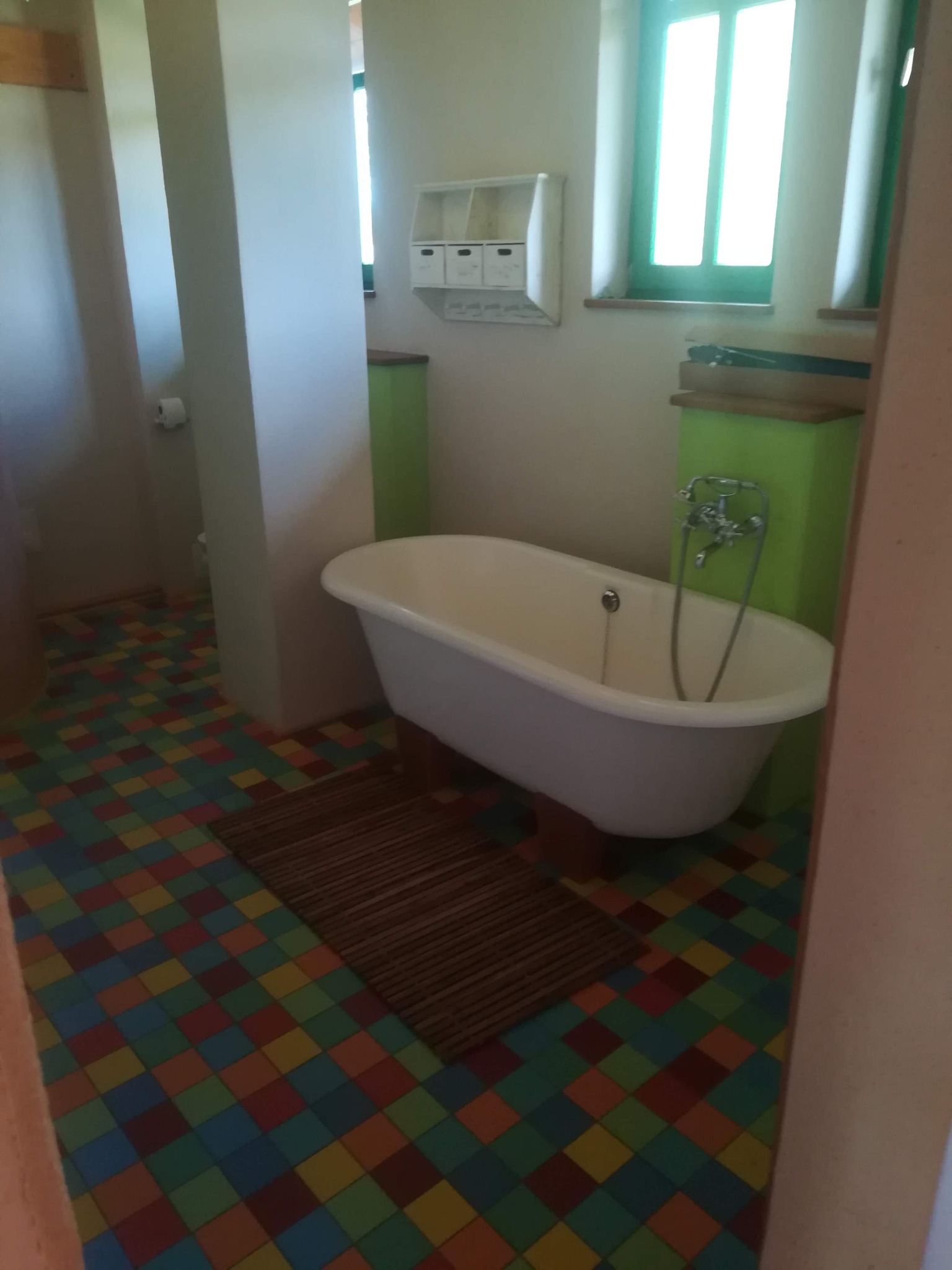 Retro łazienka z wolnostojącą wanną na nóżkach, kolorową podłogą w kratkę i zielonymi akcentami przy oknie.