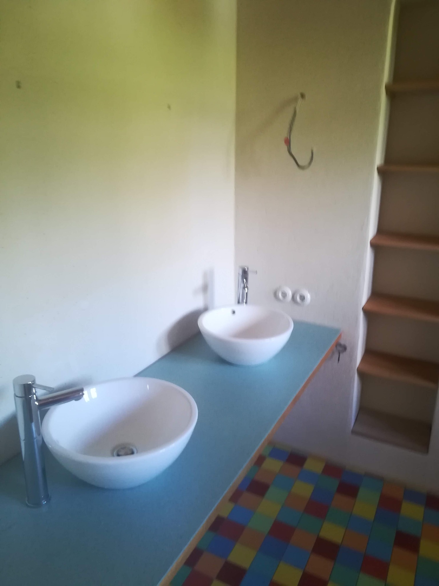 Dwie umywalki nablatowe w kształcie misy z chromowanymi bateriami na niebieskim blacie, kolorowa mozaika na podłodze, widoczny hak na ścianie i półki wnękowe.