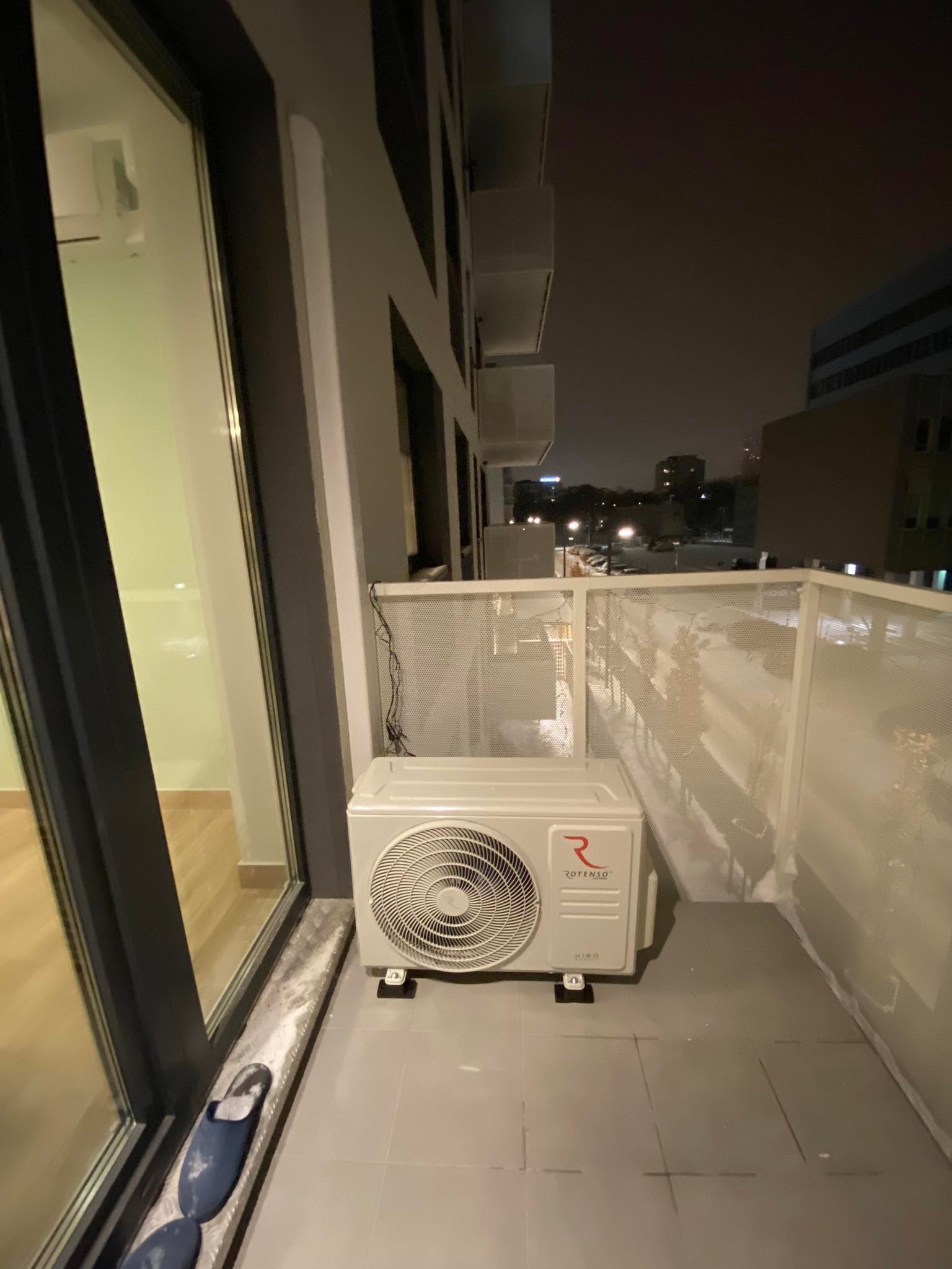 Biała jednostka zewnętrzna klimatyzacji Rotenso Hiro na balkonie apartamentu, widok na nocne miasto w tle.