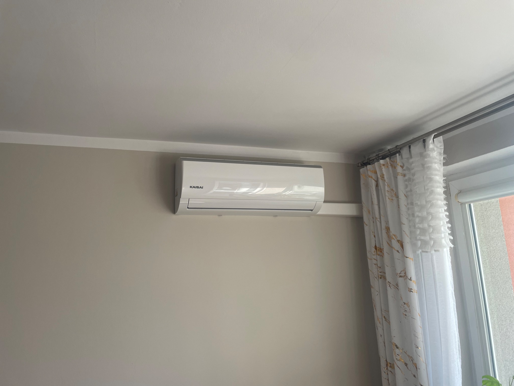 Biały klimatyzator marki Kaisai zamontowany na ścianie w jasnym pokoju, widoczne firanki w oknie.