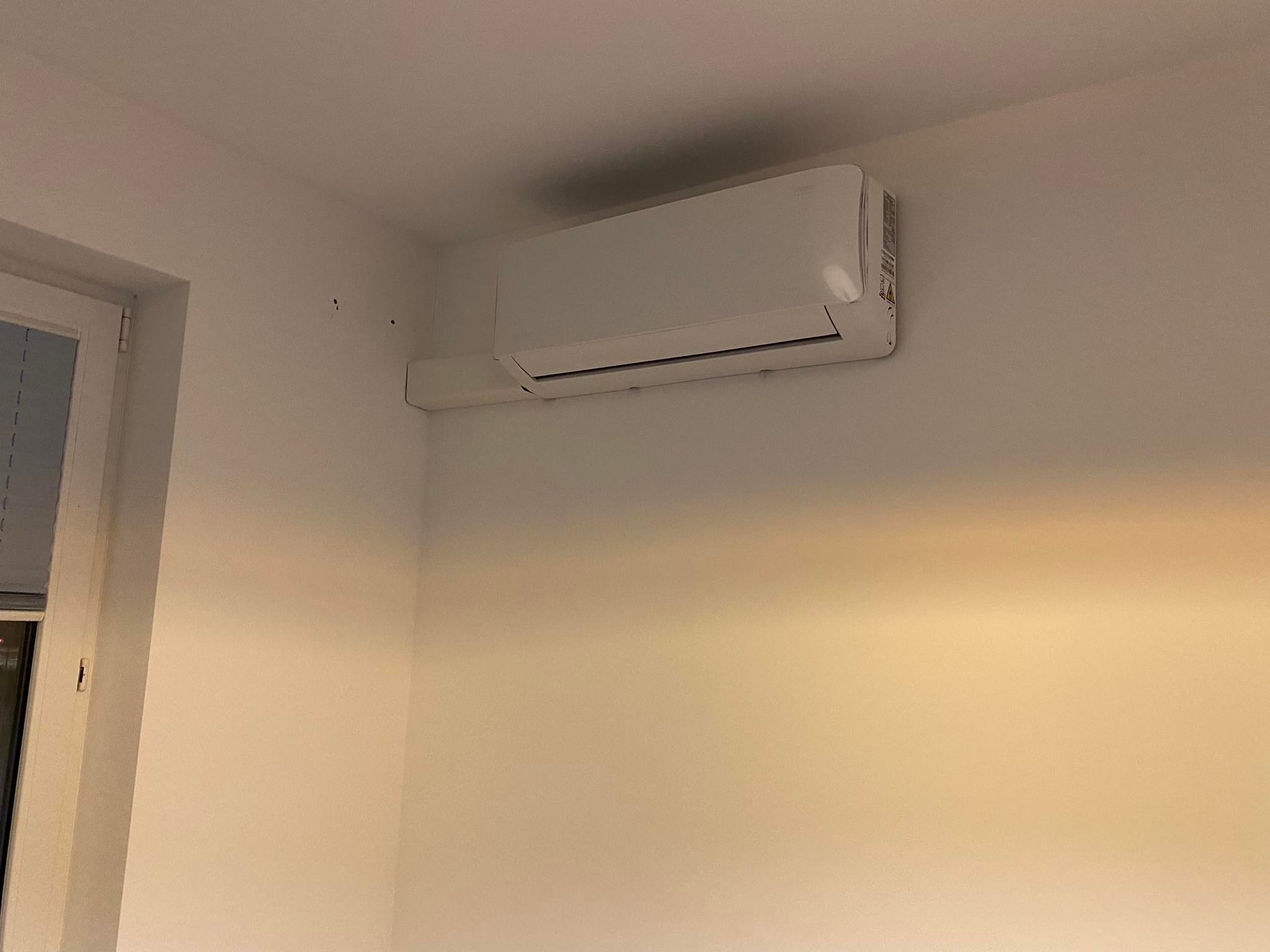 Biały klimatyzator ścienny zamontowany w rogu pokoju o białych ścianach, widoczne ślady po wierceniu.