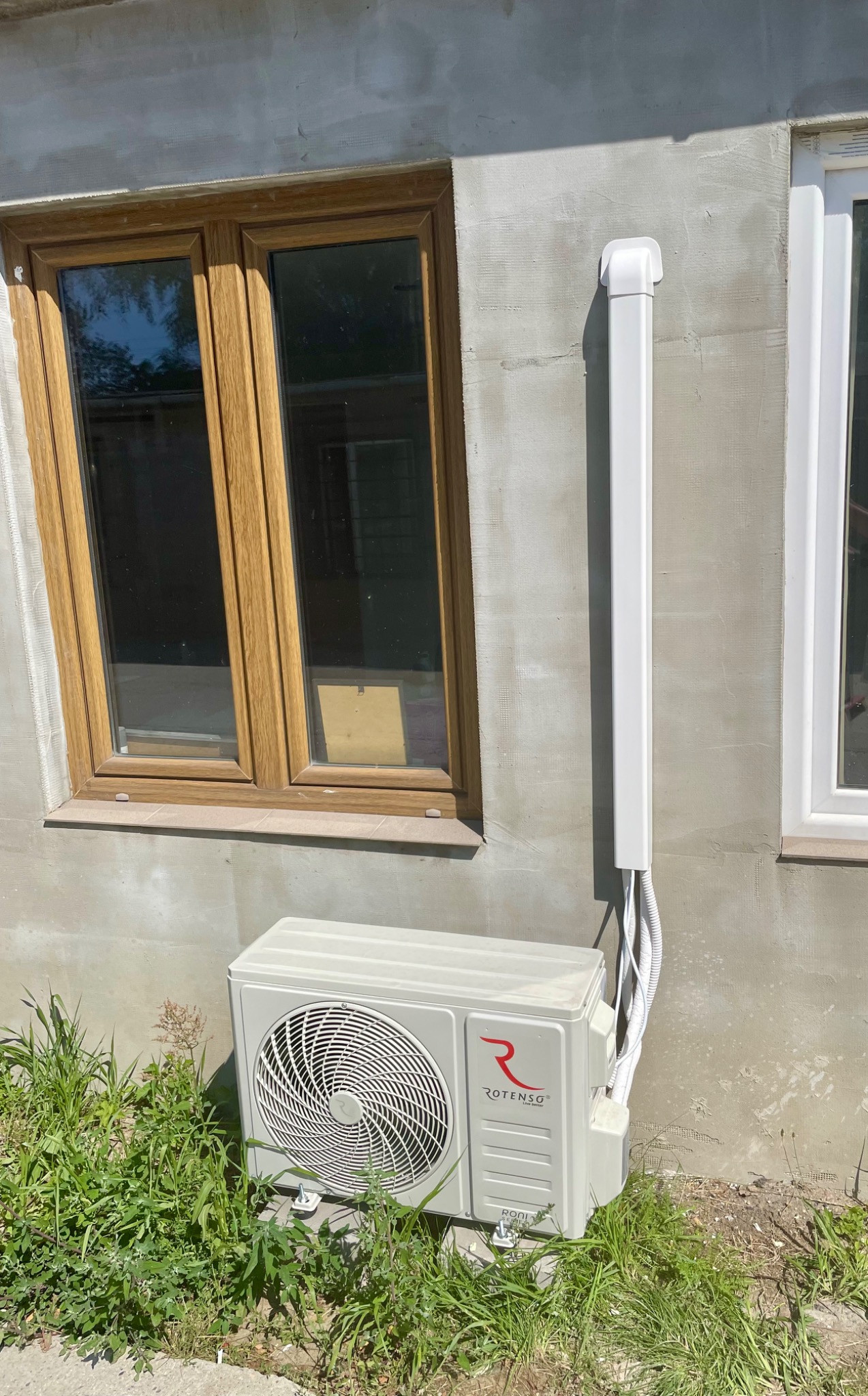Zewnętrzna jednostka klimatyzacji Rotenso zamontowana na ścianie budynku, obok okna z brązową ramą, z widocznymi przewodami w białej maskownicy i zieloną trawą u podstawy.