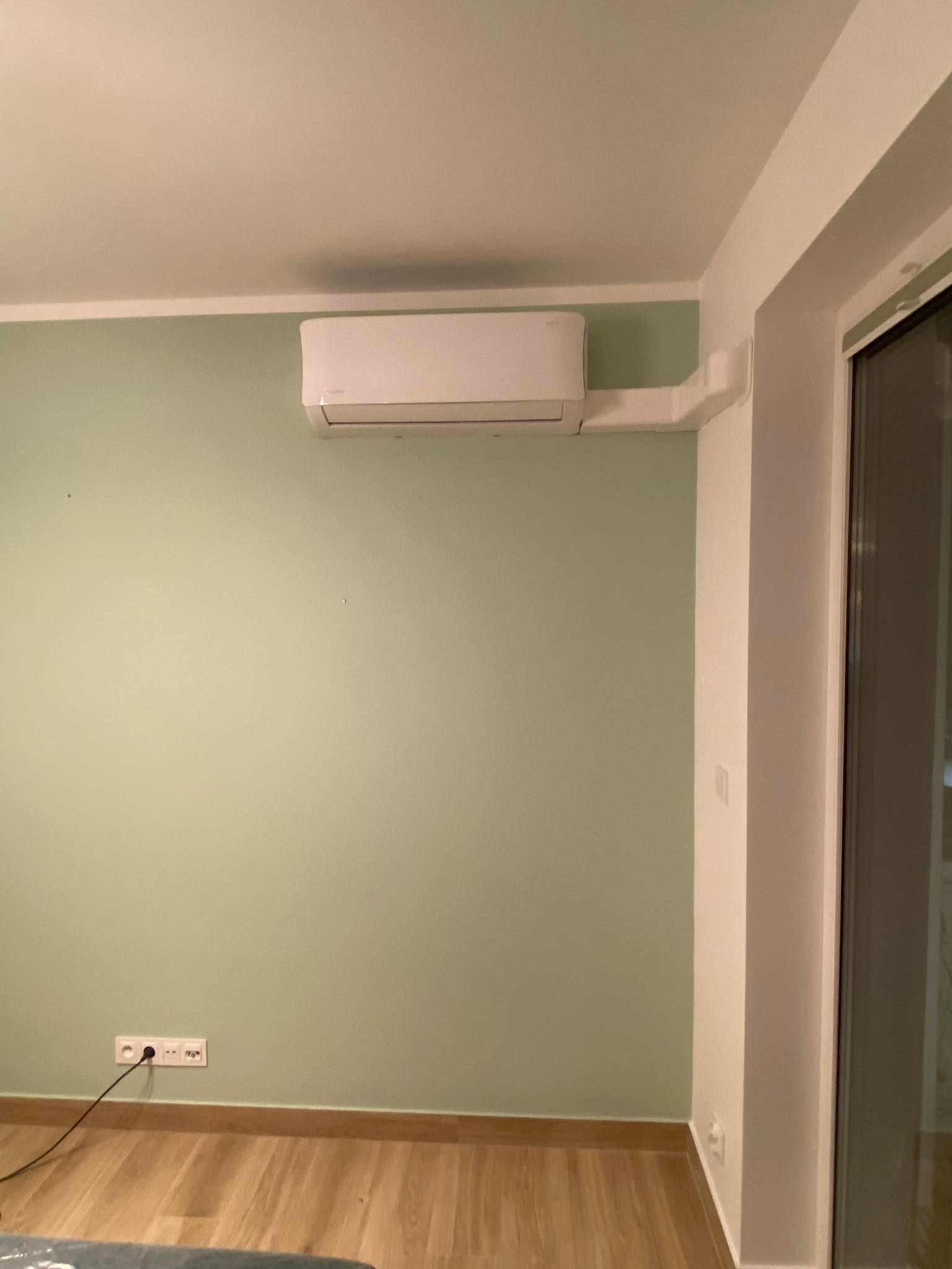 Biała jednostka klimatyzacyjna zamontowana na ścianie w pokoju o pistacjowej barwie, z widocznymi plastikowymi kanałami prowadzącymi do okna.