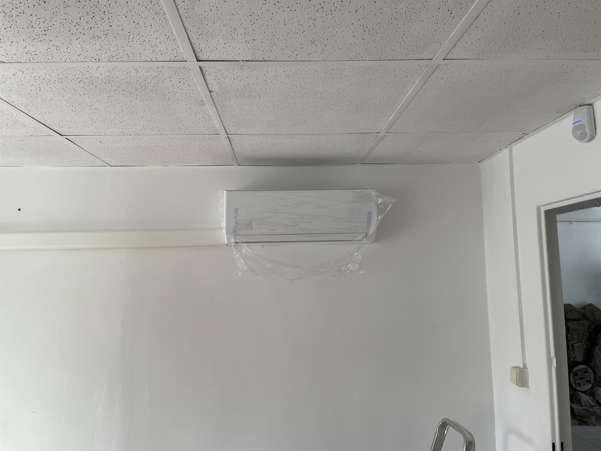 Nowo zainstalowana, biała jednostka klimatyzacyjna, zabezpieczona folią ochronną na białej ścianie w pomieszczeniu z kasetonowym sufitem.