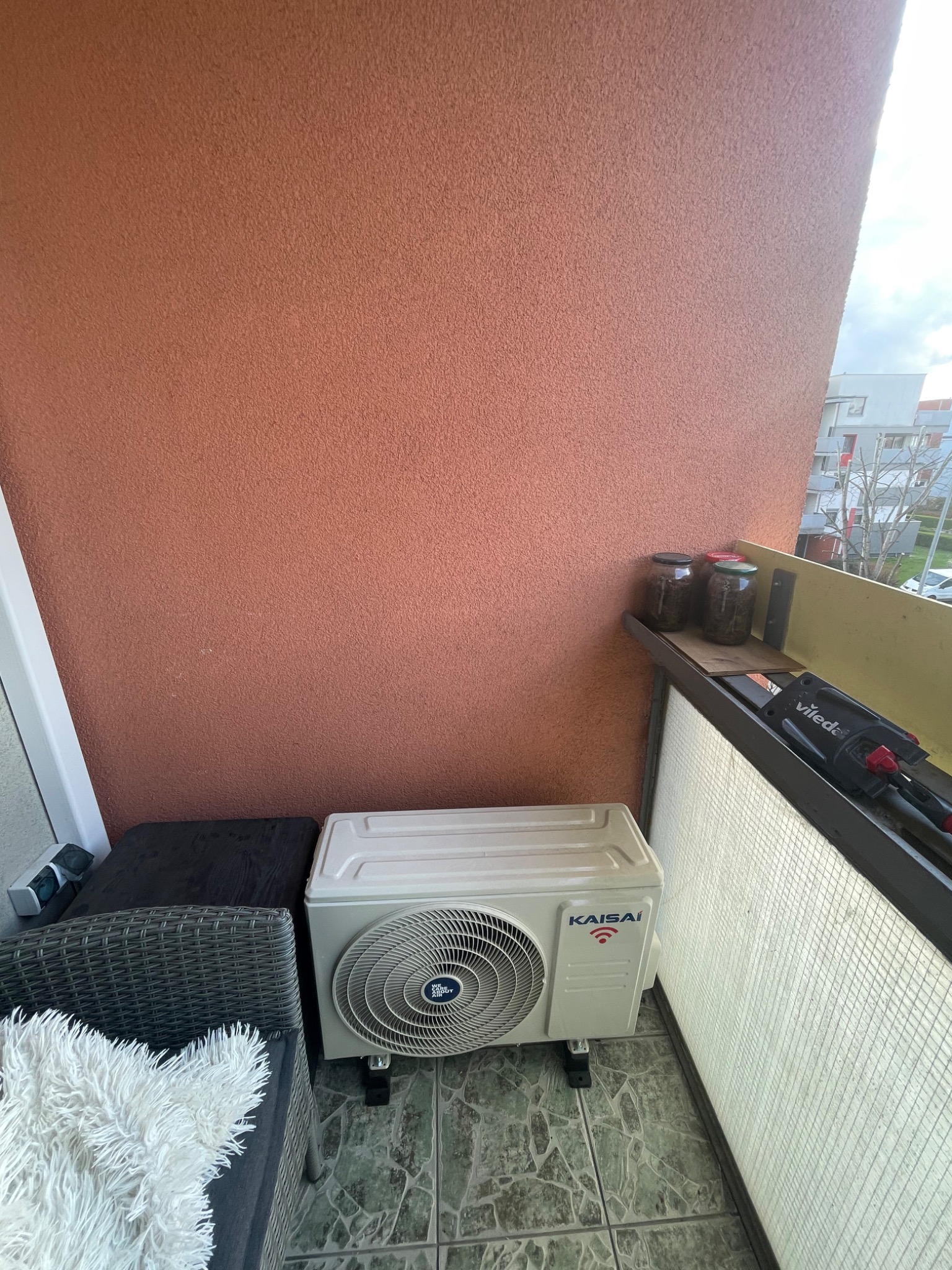 Jednostka zewnętrzna klimatyzacji Kaisai zamontowana na balkonie we Wrocławiu, obok plecionej sofy z puszystą poduszką, z widocznym fragmentem ściany w odcieniu terakoty.