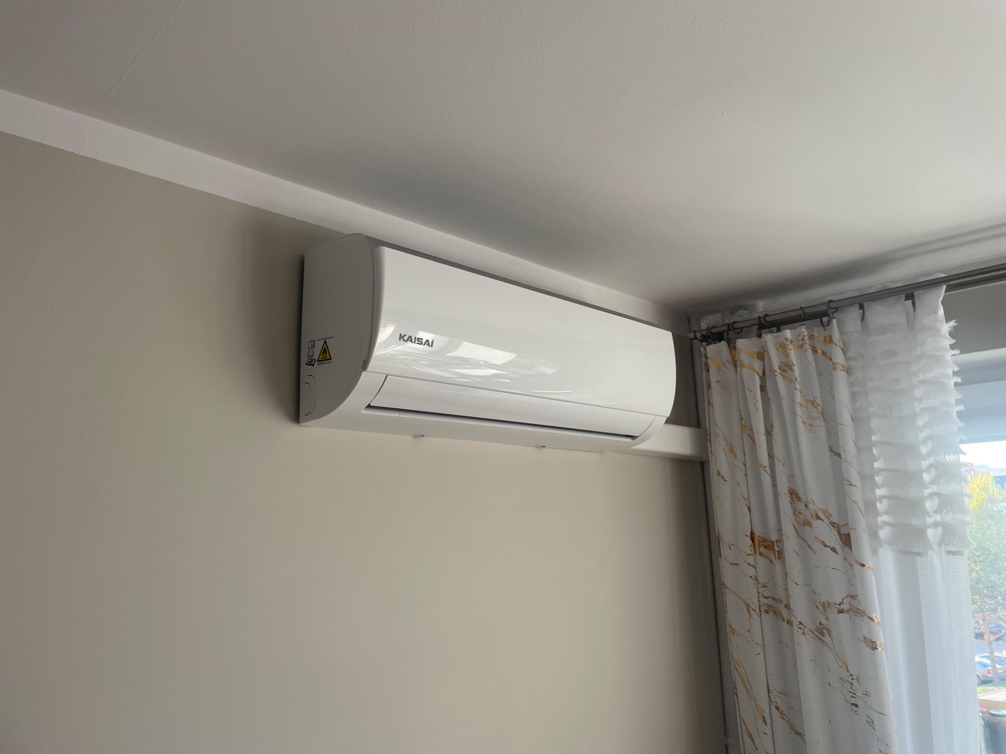 Biały klimatyzator Kaisai zamontowany na beżowej ścianie w pokoju, obok okna z białymi zasłonami w złote wzory.