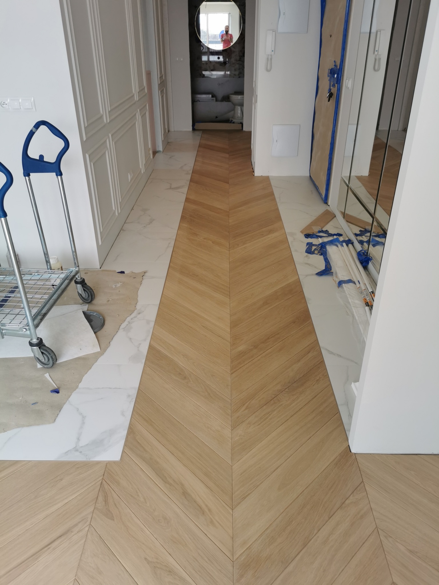 Fragment korytarza w trakcie remontu z ułożonym parkietem w jodełkę francuską, widoczne wózki transportowe i materiały zabezpieczające