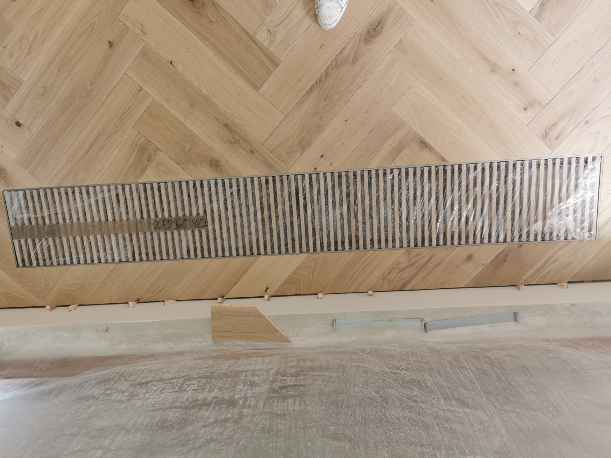 Fragment układania parkietu w jodełkę, widoczna kratka podłogowa z folią ochronną i białe buty w górnym kadrze, częściowo zakryta podłoga folią malarską.