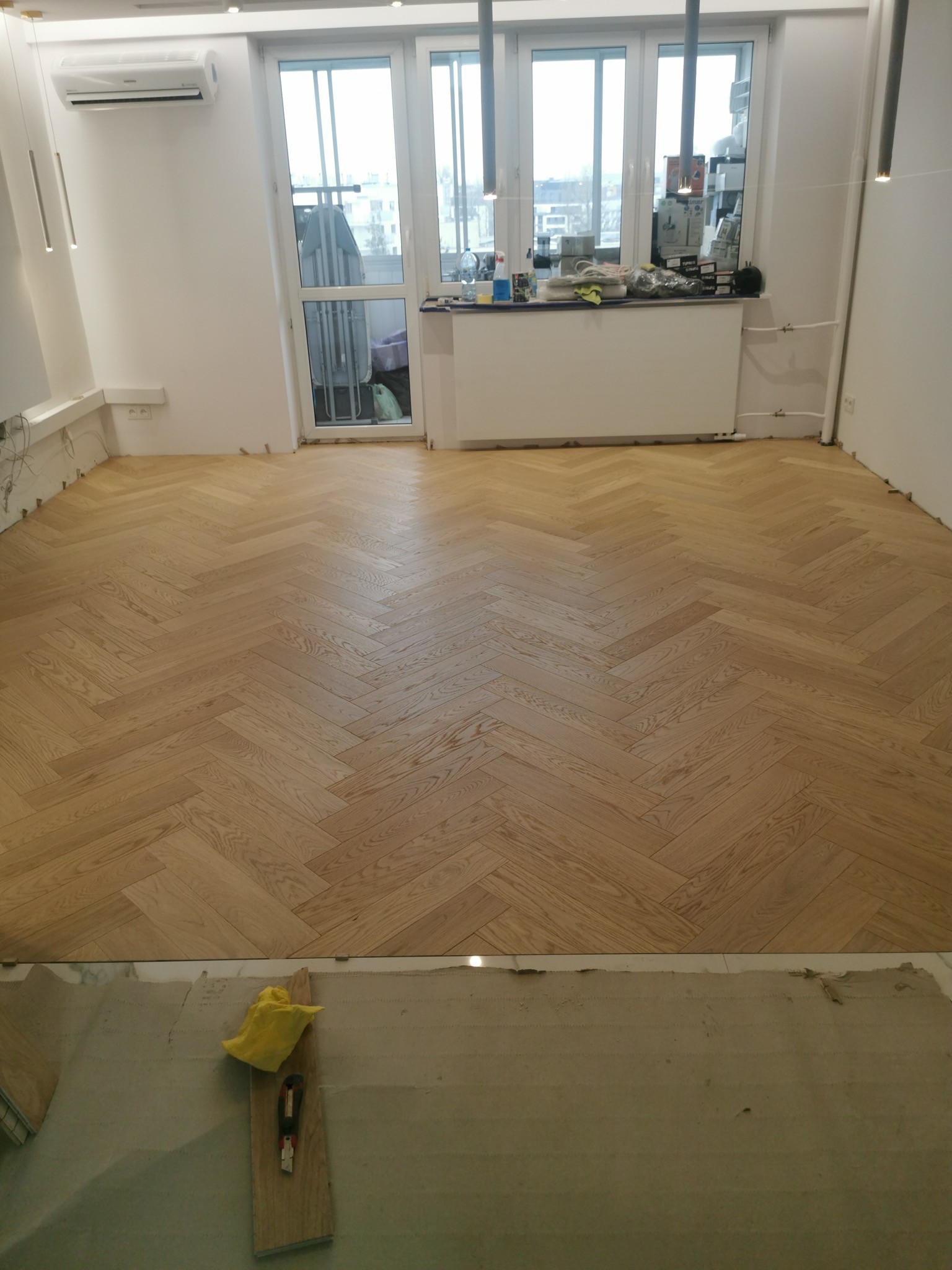 Ukończona podłoga z parkietu ułożonego w jodełkę francuską, widoczne narzędzia i żółta ściereczka na podkładzie pod parkiet, w tle okno balkonowe i białe ściany.