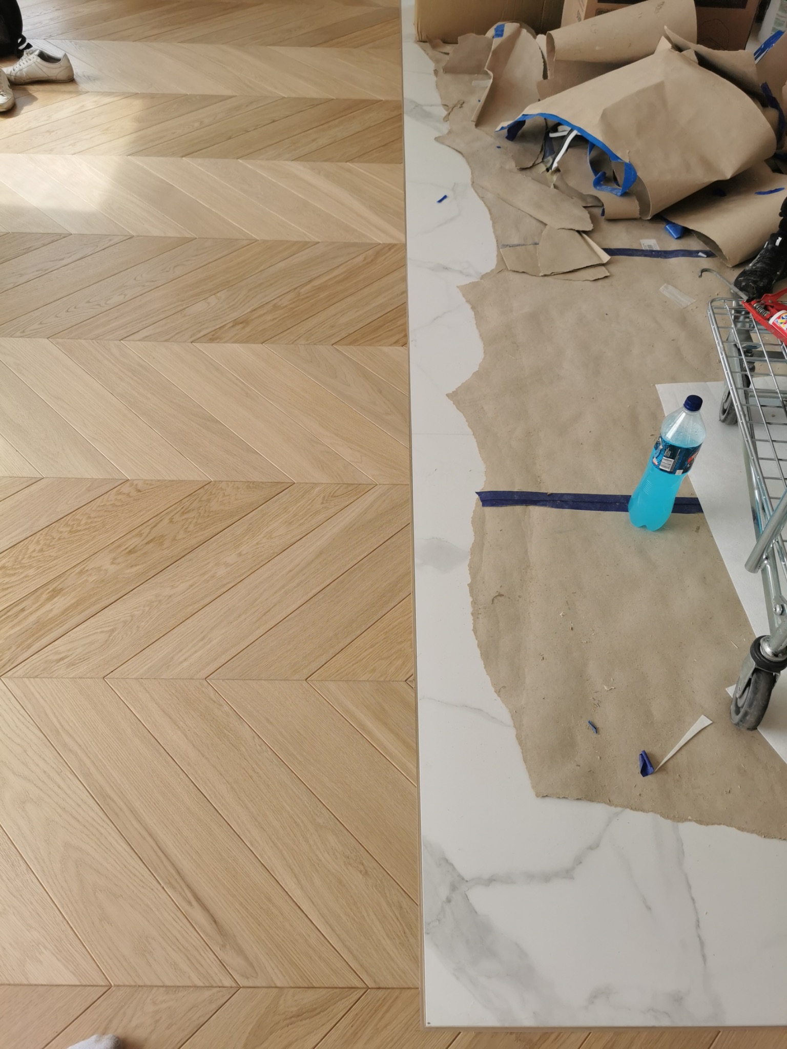 Fragment podłogi w jodełkę z jasnego drewna, świeżo położony parkiet w Otwocku, kontrastujący z zabezpieczoną folią marmurową posadzką w trakcie prac wykończeniowych.