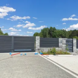 Nowoczesna, pozioma, szara brama przesuwna oraz fragment ogrodzenia z szarych paneli i słupków z szarej kostki betonowej. Na podjeździe porozrzucane narzędzia i akcesoria montażowe. Część...