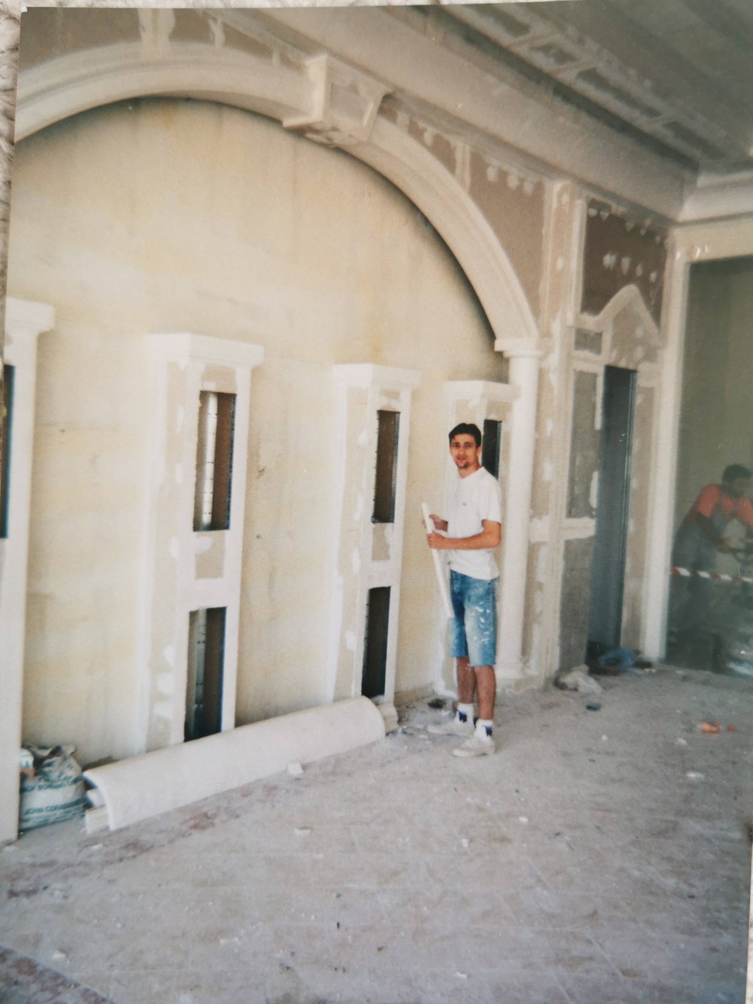 Wnętrze w trakcie prac wykończeniowych: widoczne elementy dekoracyjne ścian przygotowane do gładzi, pracownik z łatą w ręku, podłoga częściowo zakryta gruzem, w tle drugi pracownik.