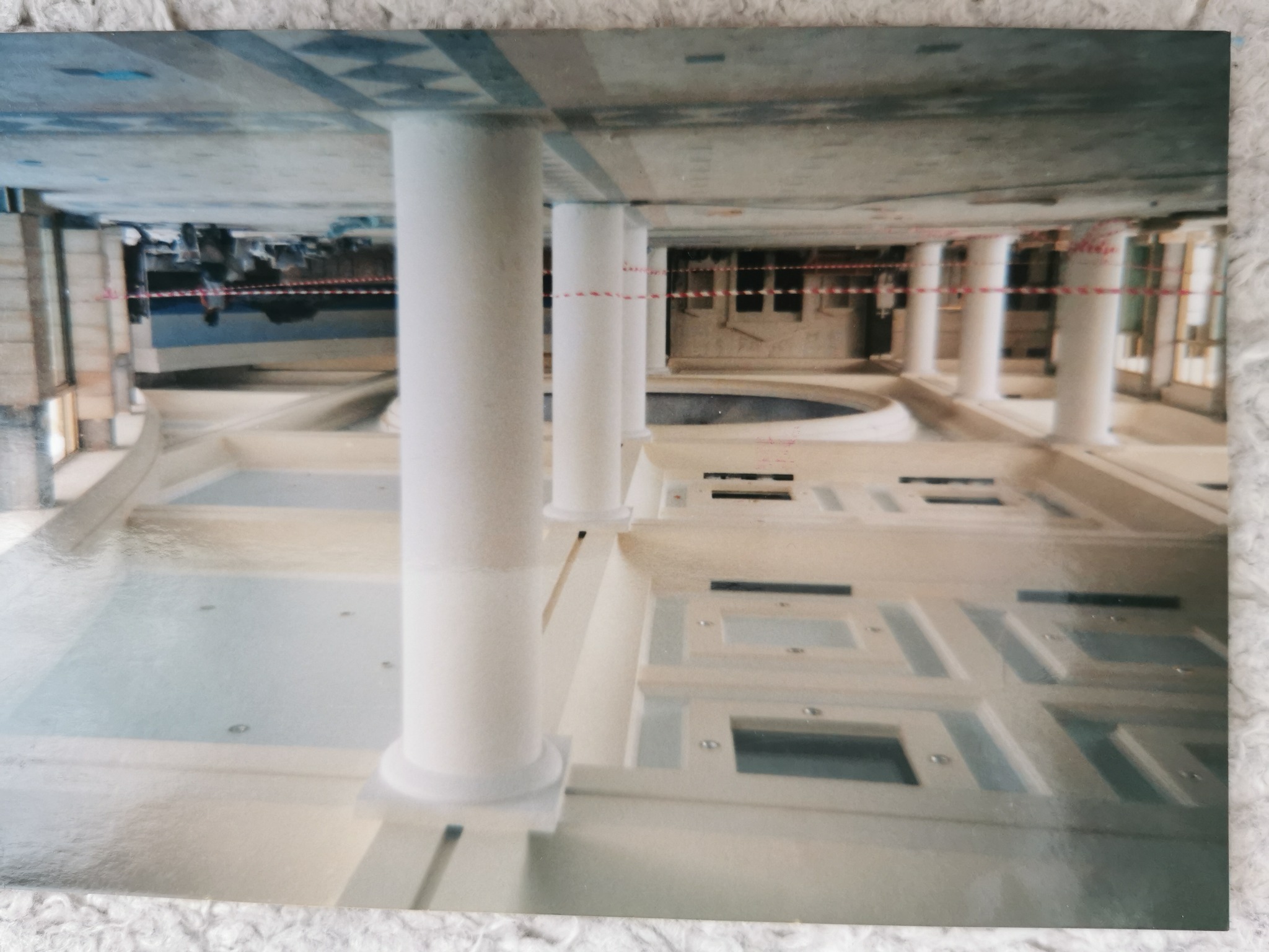 Wnętrze w trakcie prac wykończeniowych, widoczne kolumny, surowy betonowy sufit z geometrycznym wzorem, częściowo wypełnione wodą zagłębienia w podłodze, otoczone barierkami zabezpieczającymi.