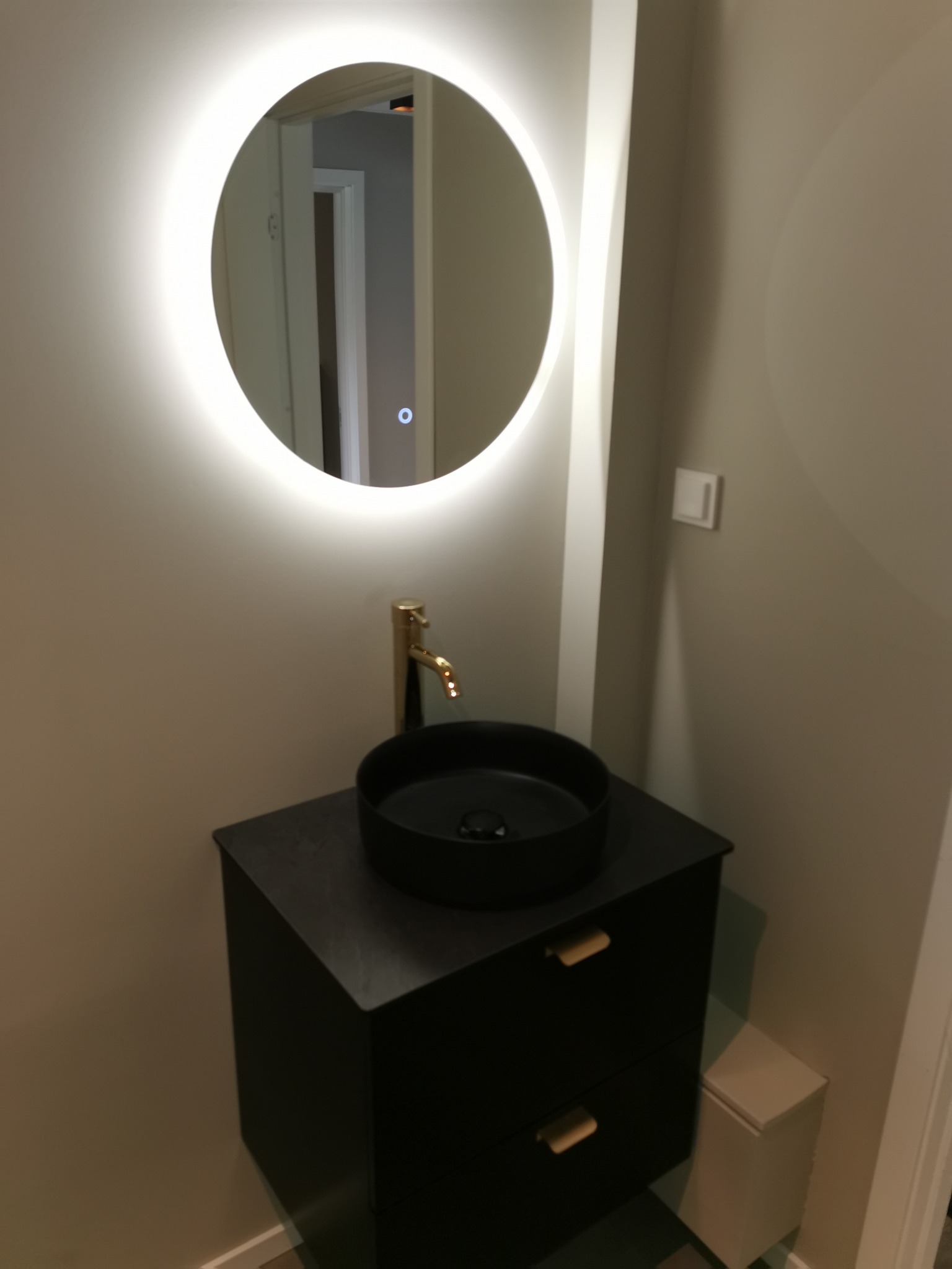 Nowoczesna łazienka z czarną umywalką nablatową, złotą baterią i okrągłym lustrem z podświetleniem LED, umieszczona na szafce z dwiema szufladami i drewnianymi uchwytami.