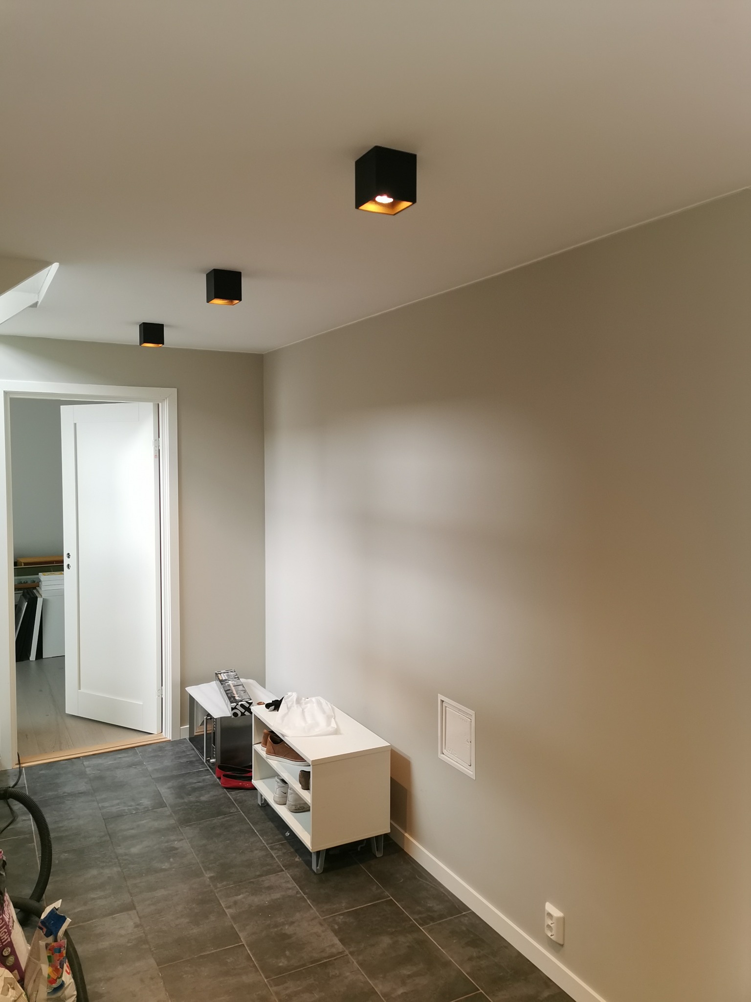 Wnętrze pokoju z szarymi ścianami, ciemną podłogą z płytek, trzema czarnymi, kwadratowymi lampami sufitowymi, białym regałem na buty i otwartymi drzwiami do kolejnego pomieszczenia.
