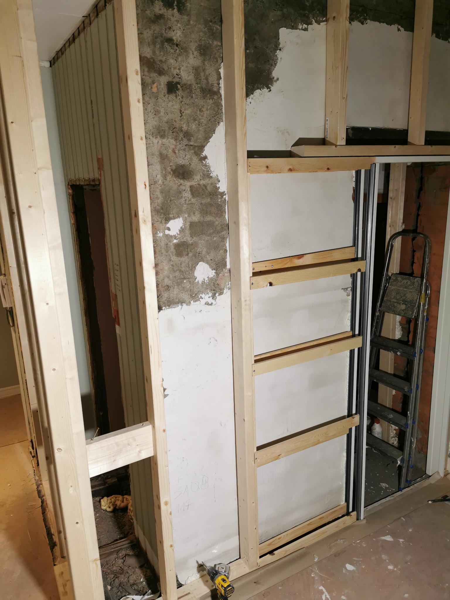 Wnętrze w trakcie prac: drewniana konstrukcja szkieletowa ścianki działowej, fragmenty starej ściany z cegły i resztkami tynku, drabina oparta o ścianę, widoczne narzędzia pracy.