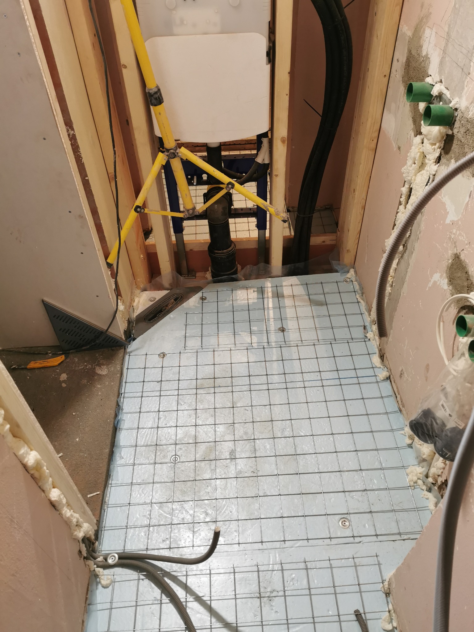 Przygotowanie podłoża pod posadzkę w łazience: siatka zbrojeniowa na folii, widoczne rury i odpływ liniowy, ściany z pianką montażową.