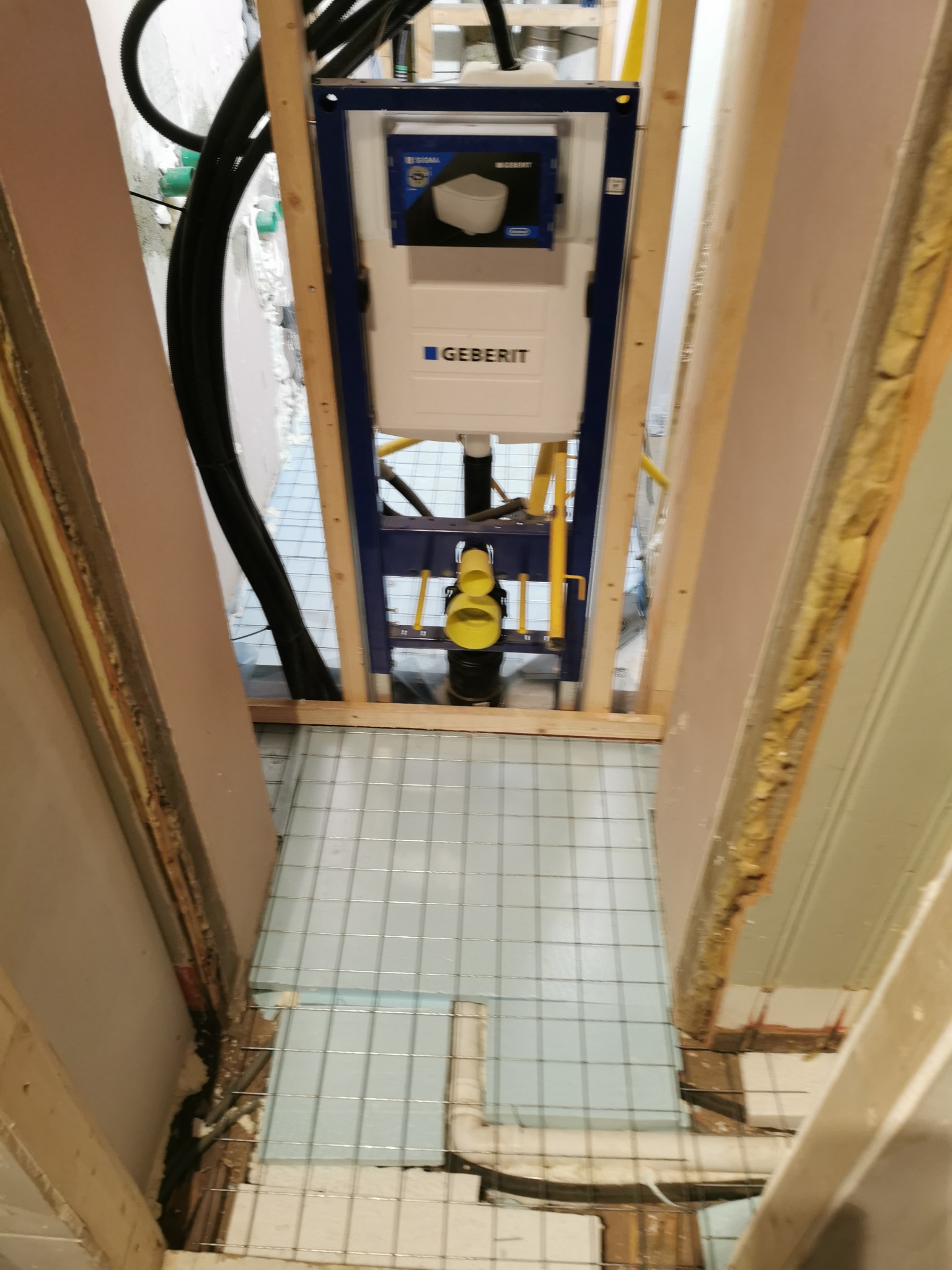 Instalacja podtynkowa Geberit w trakcie montażu w łazience, widoczne rury doprowadzające wodę, stelaż obudowany drewnianą konstrukcją, fragment podłogi z płytkami i siatką wzmacniającą.