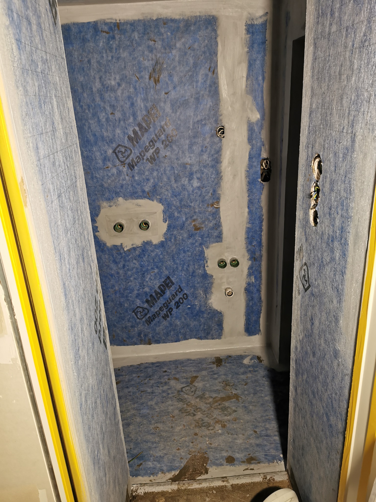 Przygotowanie podłoża w łazience: hydroizolacja ścian i podłogi z widocznymi puszkami elektrycznymi, zabezpieczonymi taśmą. Widoczny fragment żółtej taśmy zabezpieczającej przy drzwiach.