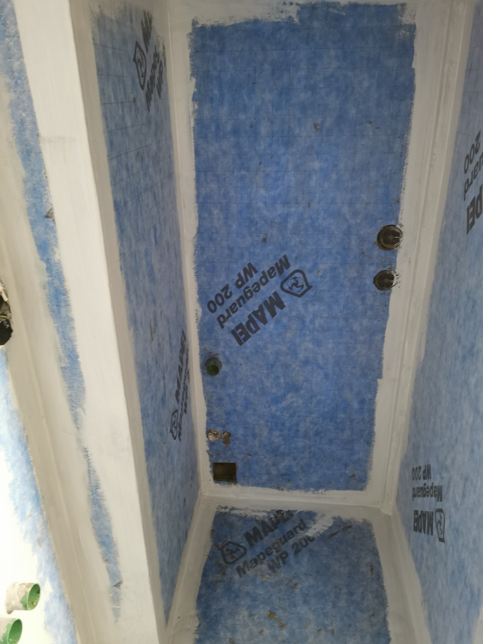 Przygotowanie podłoża w łazience, wyłożone niebieską membraną hydroizolacyjną Mapguard WP 200, uszczelnione białą masą w narożnikach i wokół rur, widoczne otwory na instalacje.