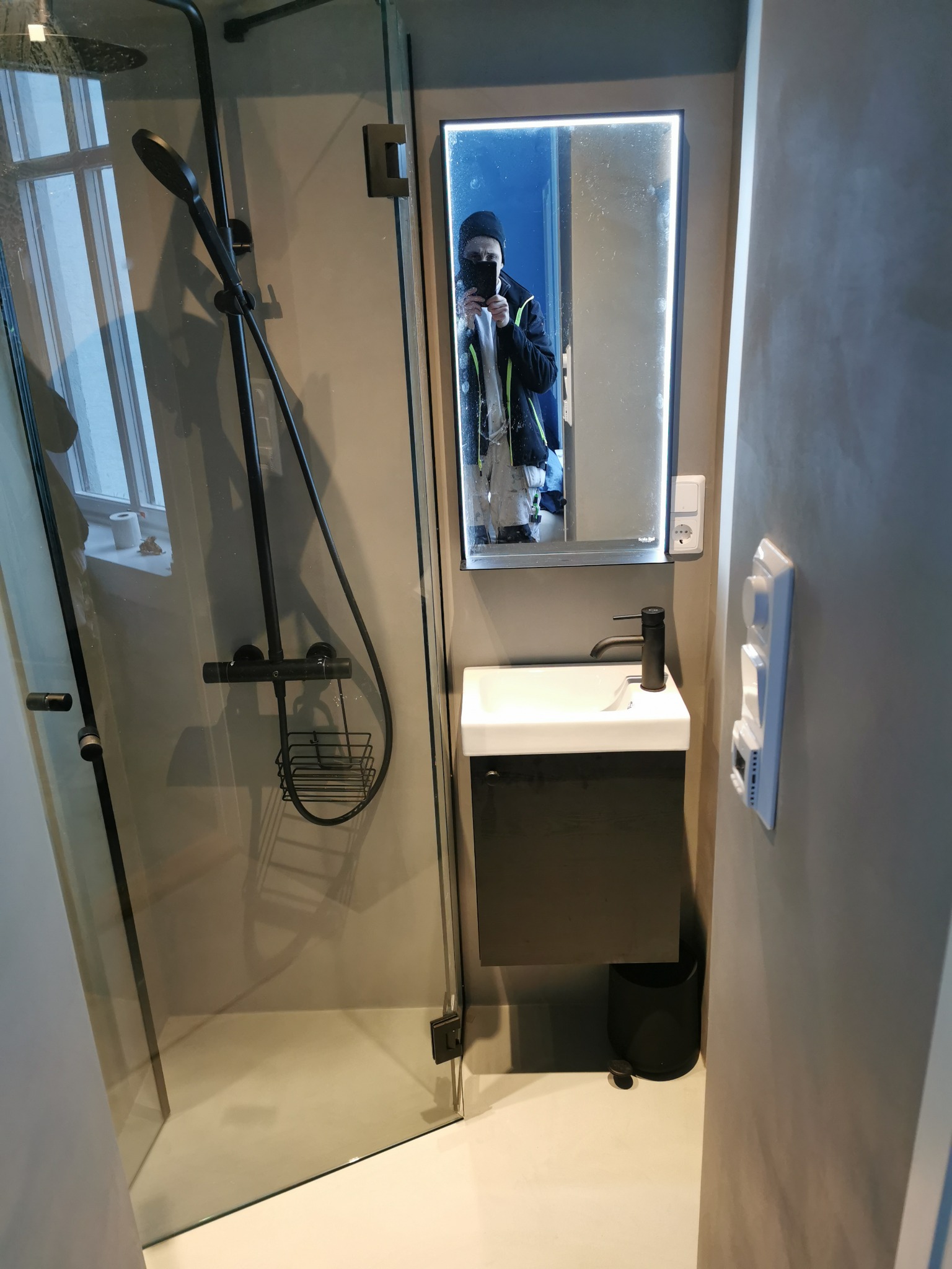 Nowoczesna łazienka z kabiną prysznicową typu walk-in, czarną armaturą i podświetlanym lustrem, w którym odbija się osoba robiąca zdjęcie.