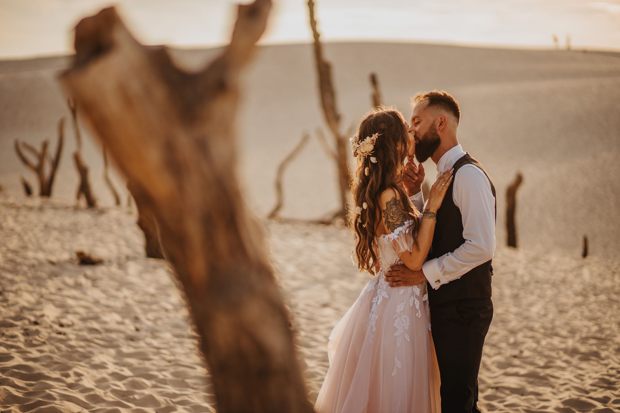 Para młoda całuje się na tle piaszczystych wydm w ciepłym, zachodzącym słońcu; panna młoda w sukni z kwiatowymi aplikacjami i wiankiem, pan młody w kamizelce i białej koszuli, pień drzewa...