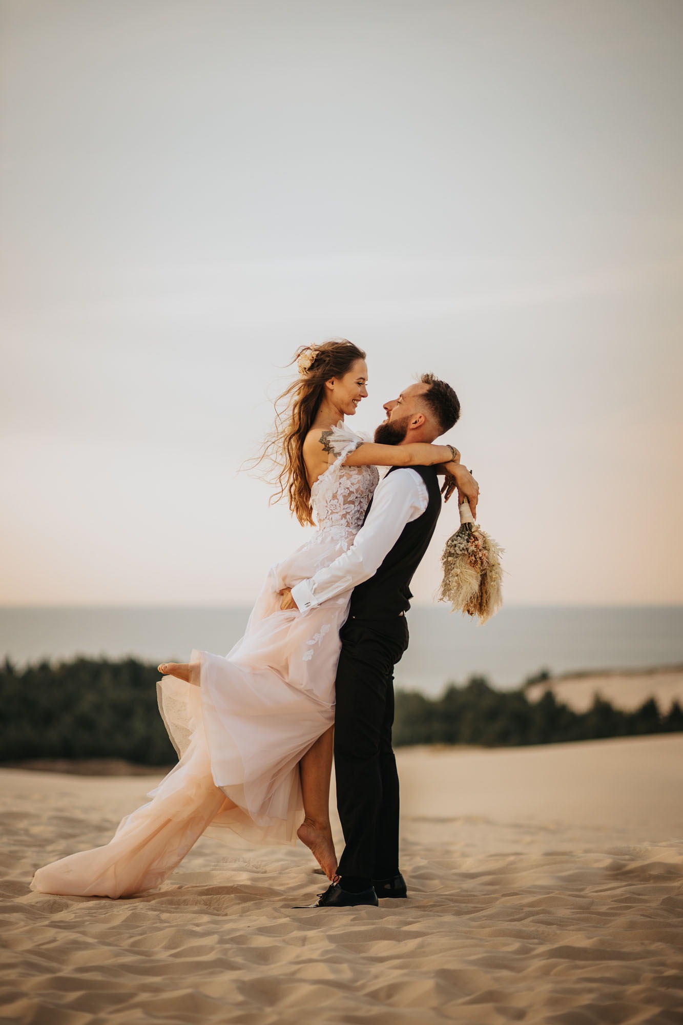 Para młoda na wydmie: panna młoda w koronkowej sukni z pudrowo-różowym tiulem, unoszona przez pana młodego w kamizelce, w tle piaszczysty krajobraz i linia lasu.