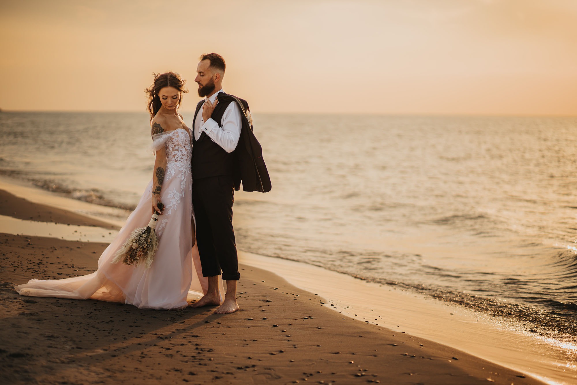 Para młoda na plaży o zachodzie słońca: panna młoda w zwiewnej sukni z koronkowymi aplikacjami i bukietem suszonych kwiatów, pan młody w garniturze z marynarką przewieszoną przez ramię, oboje boso...