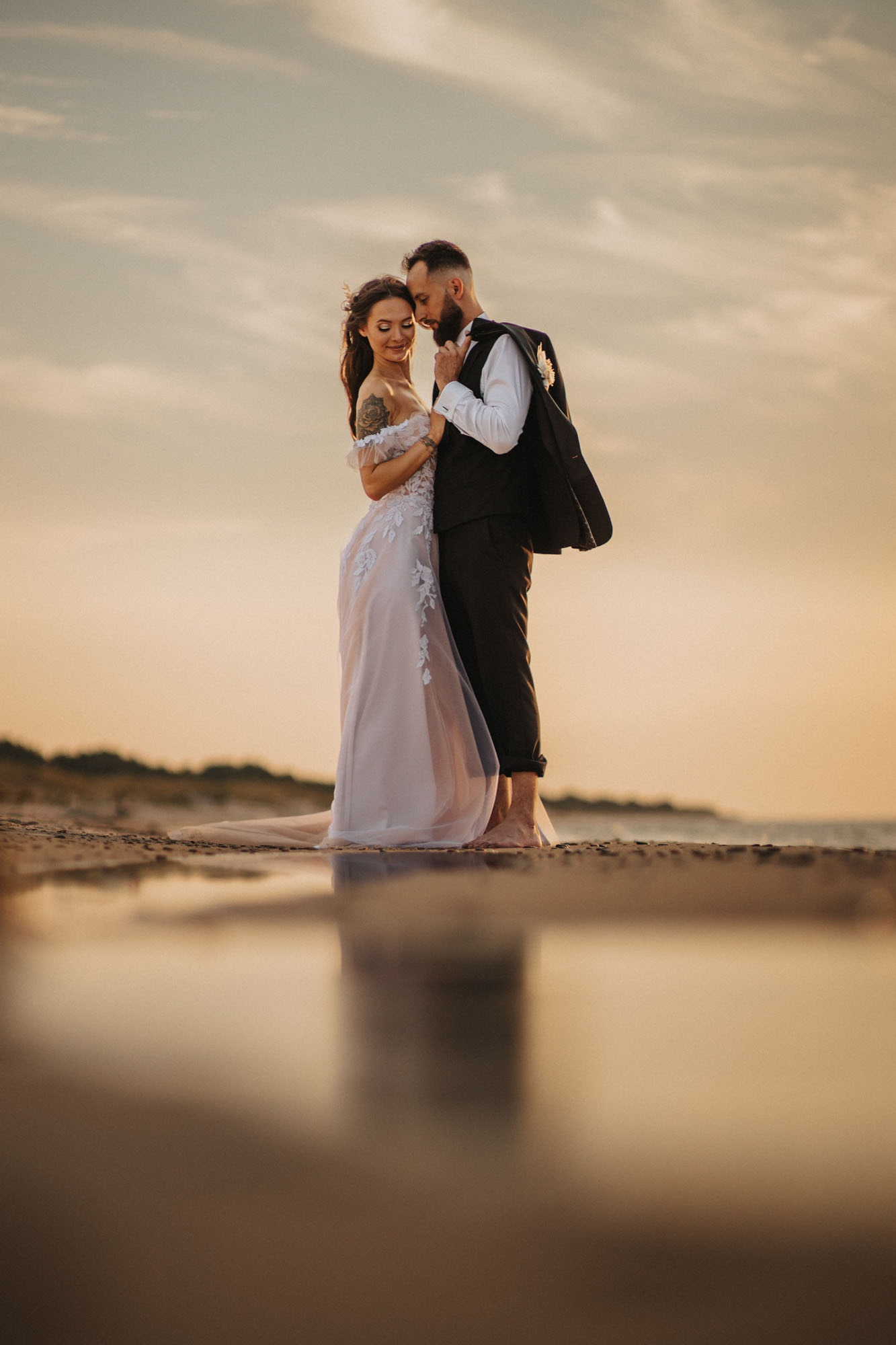 Para młoda na plaży o zachodzie słońca; panna młoda w delikatnej sukni z koronkowymi aplikacjami, pan młody w ciemnym garniturze z marynarką nonszalancko zarzuconą na ramieniu, boso na piasku.