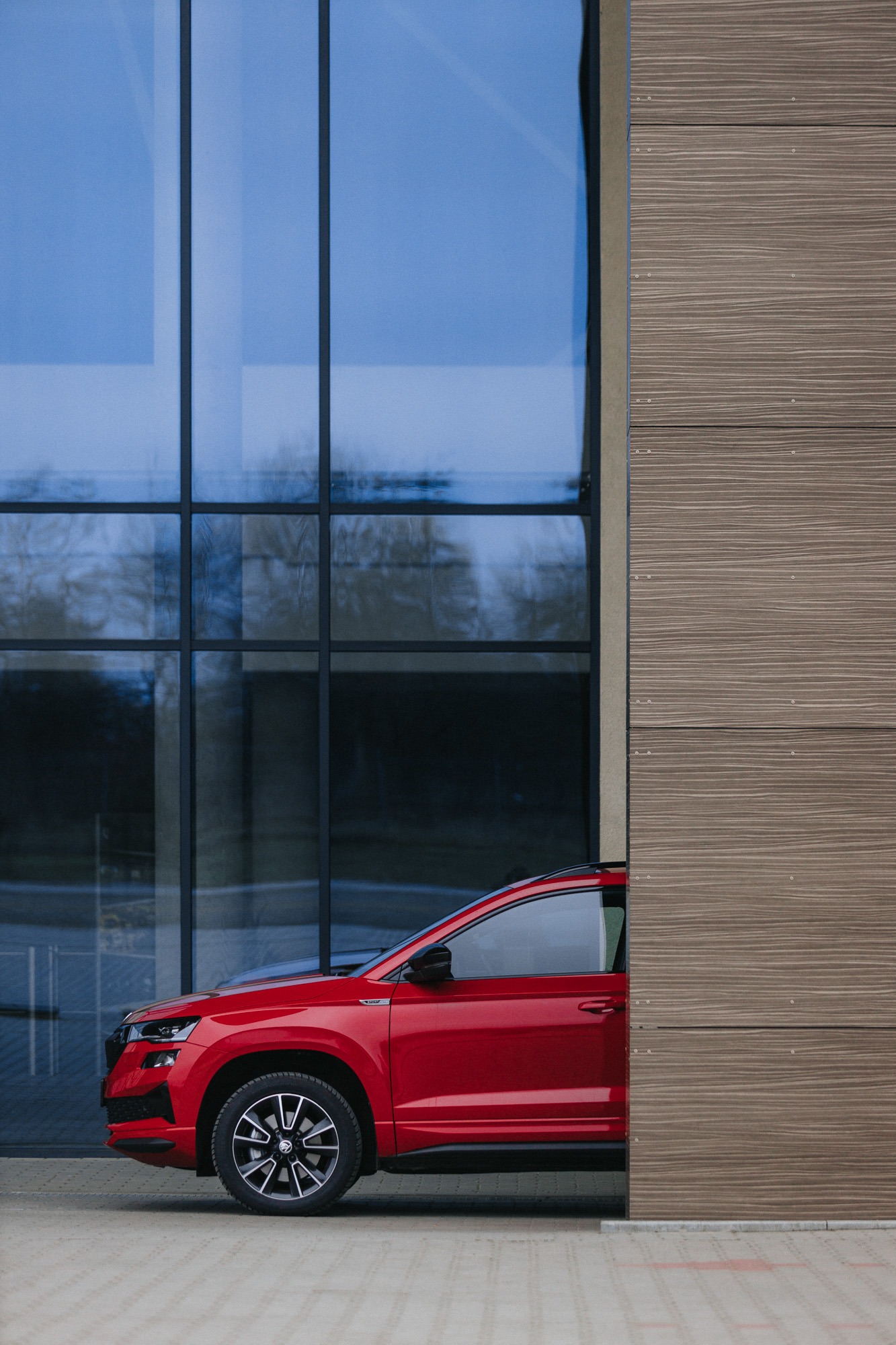 Czerwony samochód marki Skoda Karoq zaparkowany przed nowoczesnym budynkiem z szklaną fasadą i drewnianą okładziną.