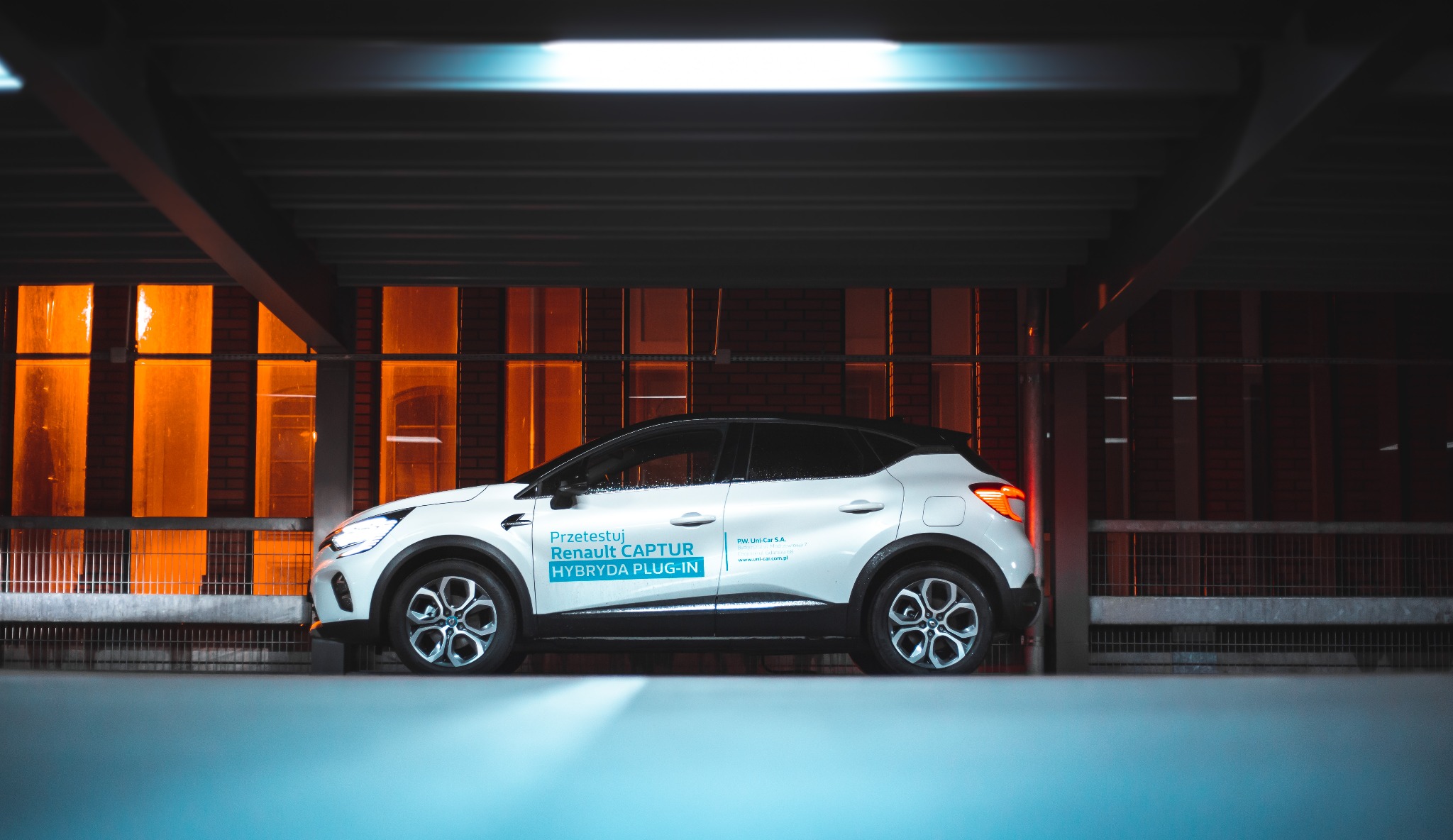 Biały Renault Captur Hybryda Plug-in zaparkowany w garażu podziemnym, widok z boku, z pomarańczowym oświetleniem w tle.