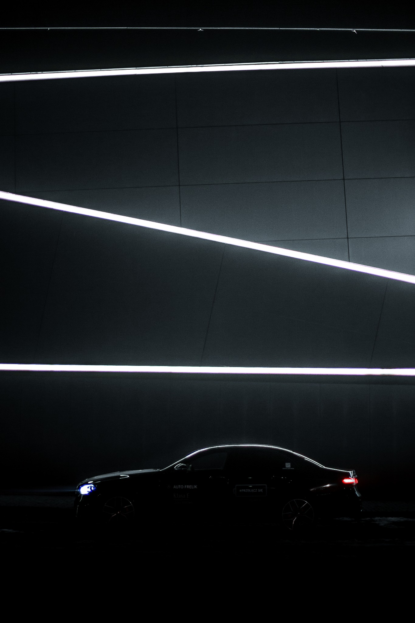 Ciemny samochód osobowy, widoczny z boku, podświetlony poziomymi liniami neonowymi na tle szarej ściany.