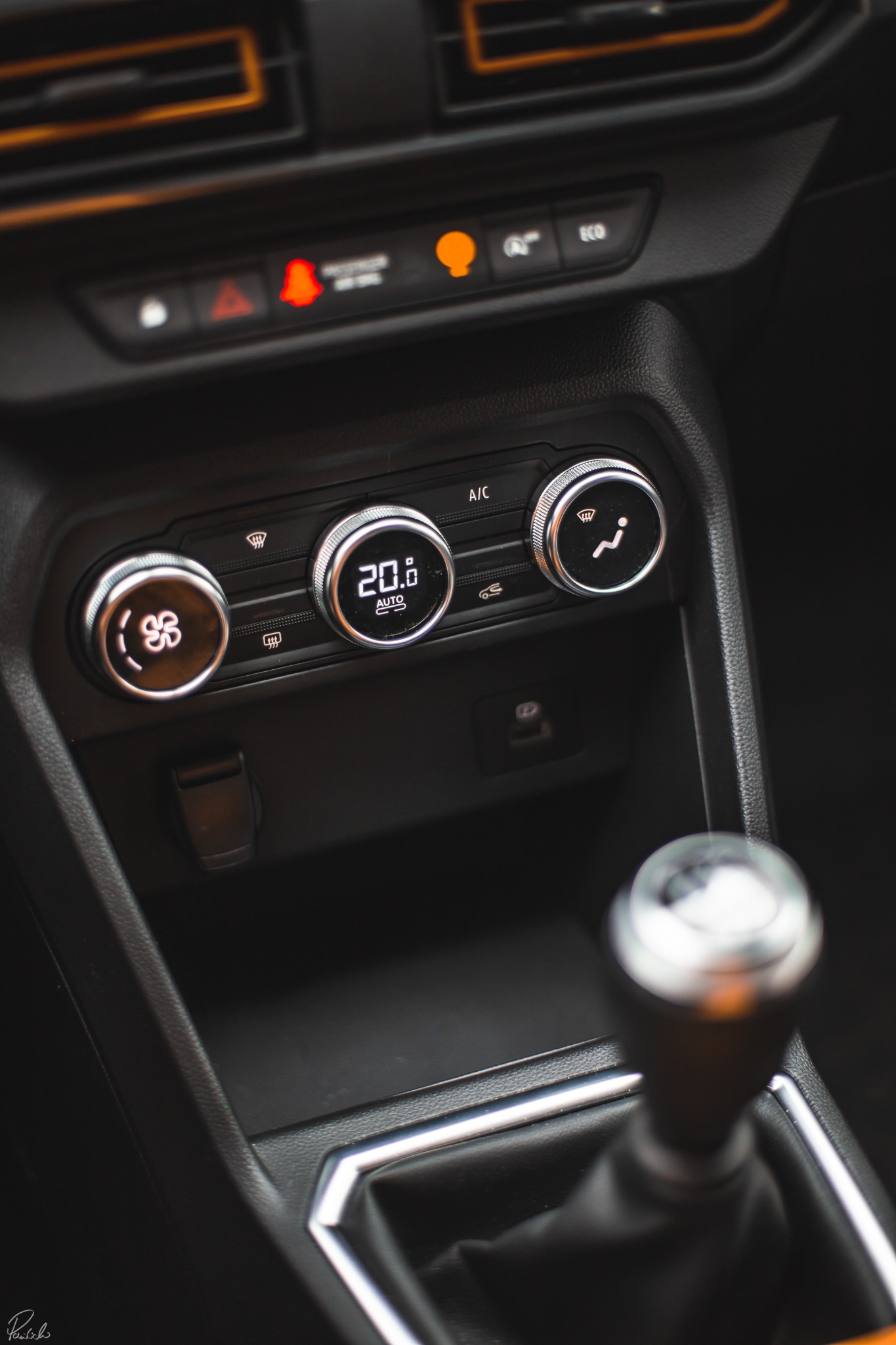 Wnętrze samochodu z bliska, skupienie na panelu klimatyzacji z wyświetlaczem 20 stopni Celsjusza, pokrętła regulacji i dźwignia zmiany biegów na pierwszym planie.
