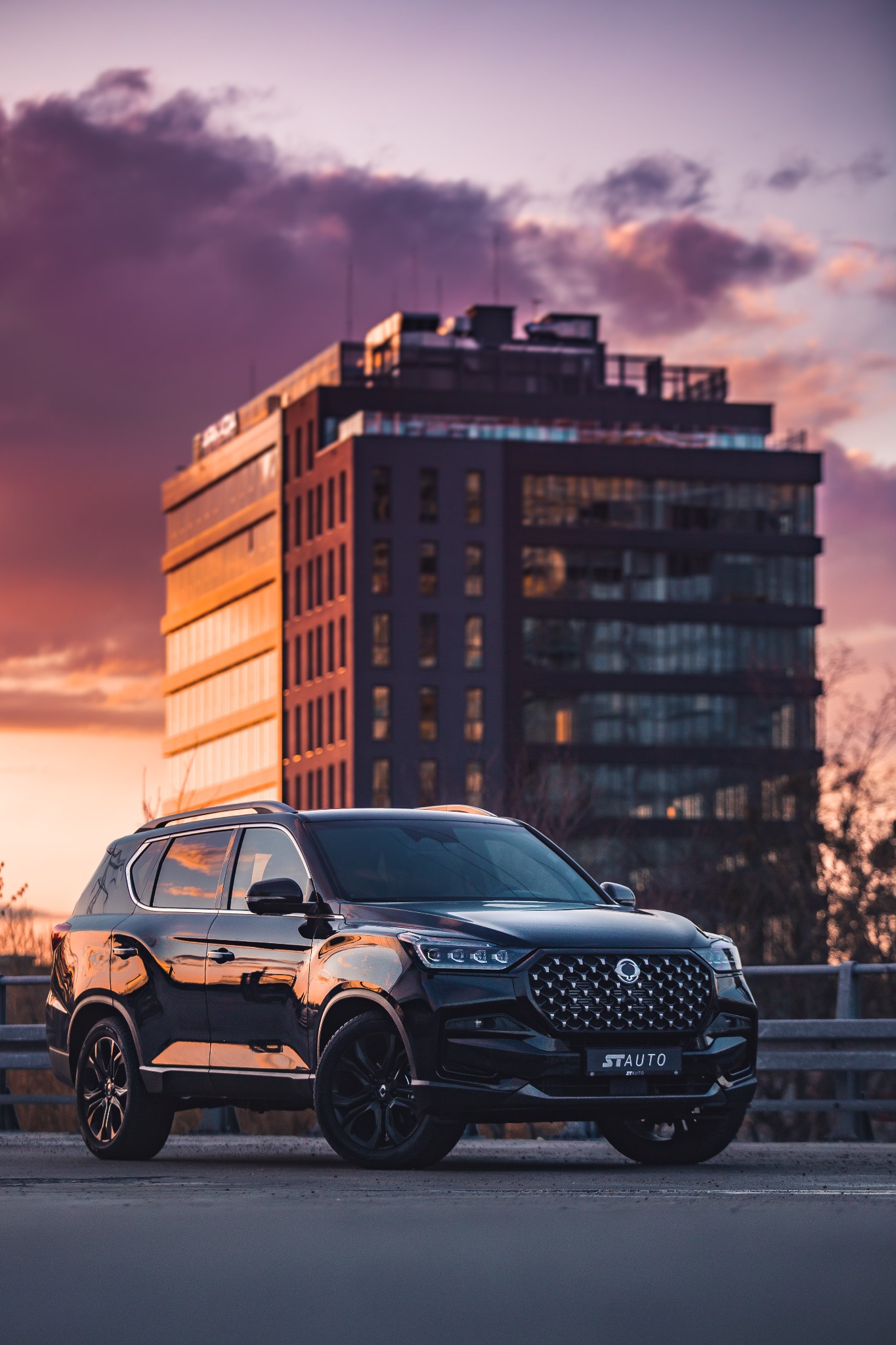Czarny SUV SsangYong Rexton stoi na tle nowoczesnego budynku biurowego podczas zachodu słońca, z widocznym logo dealera 'STAUTO' na grillu.