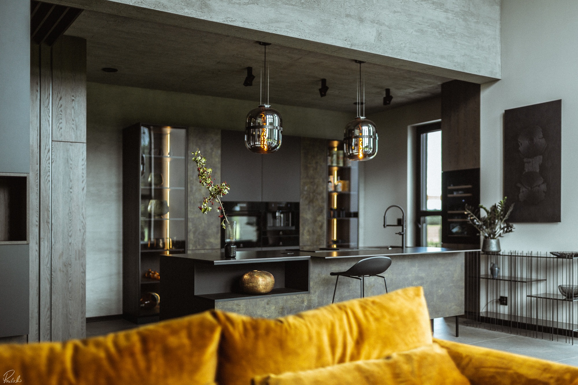 Nowoczesna kuchnia z wyspą, oświetlona wiszącymi lampami w kształcie kul, z elementami dekoracyjnymi, takimi jak złota kula i gałązka w wazonie, widziana z perspektywy żółtej sofy.