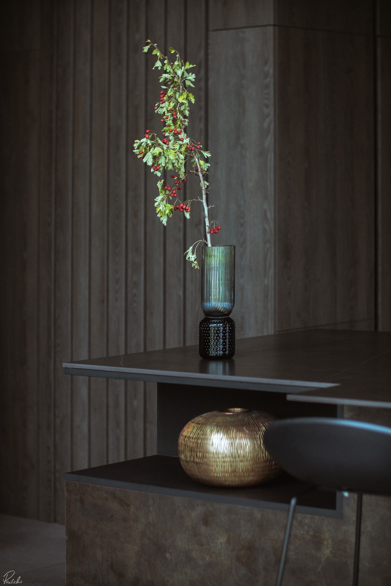 Minimalistyczna aranżacja wnętrza: gałązka jarzębiny w szklanym wazonie na czarnym blacie, złota kula dekoracyjna w tle, ciemne drewniane wykończenie.