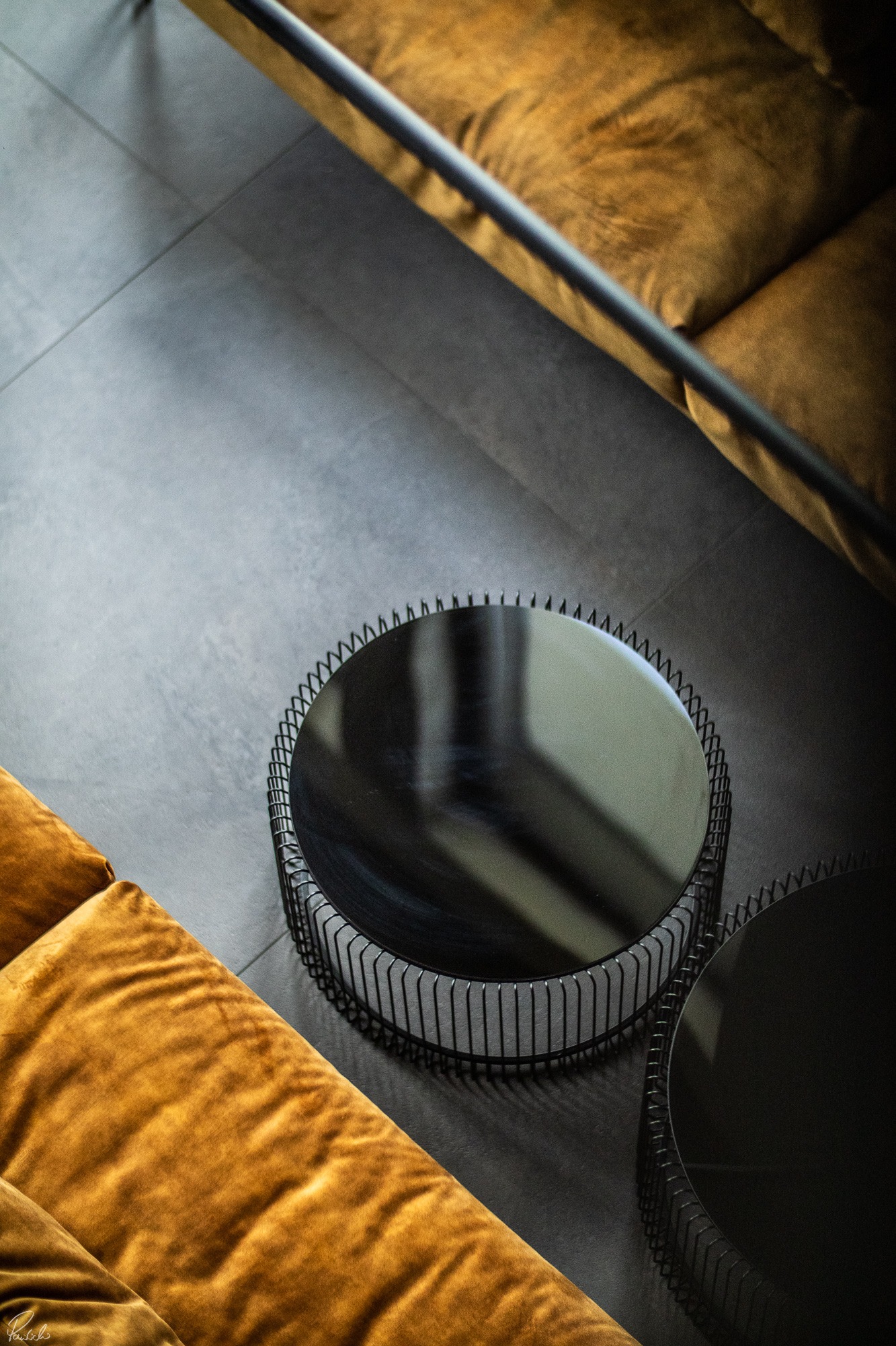 Nowoczesny salon z dwiema okrągłymi, czarnymi stolikami kawowymi z metalową konstrukcją i lustrzanym blatem, obok złotej, welurowej sofy na szarych płytkach podłogowych, widok z góry.