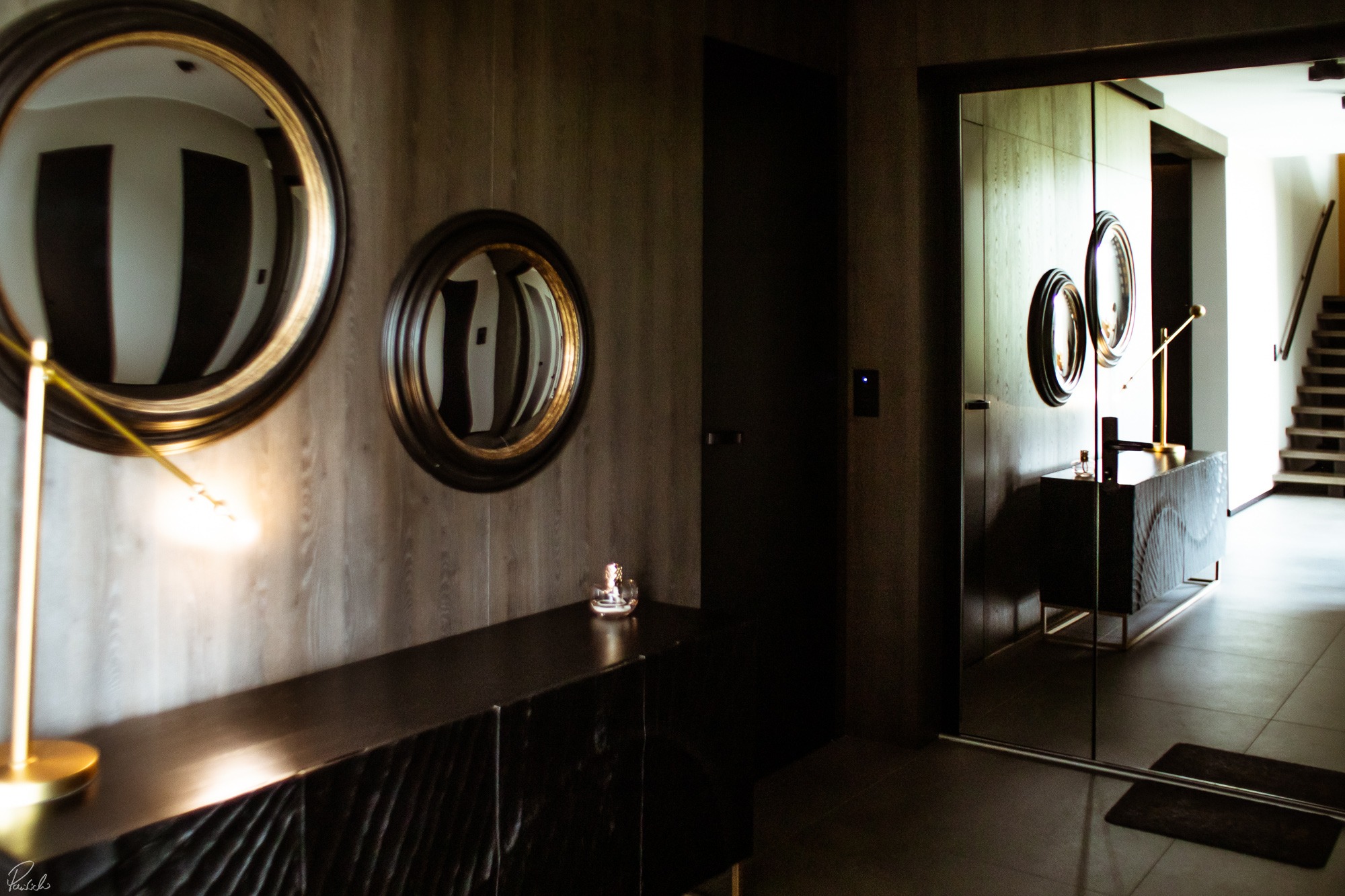 Nowoczesny przedpokój z drewnianą komodą, okrągłymi lustrami na ścianie i szklanymi drzwiami odbijającymi wnętrze, oświetlony złotą lampą.